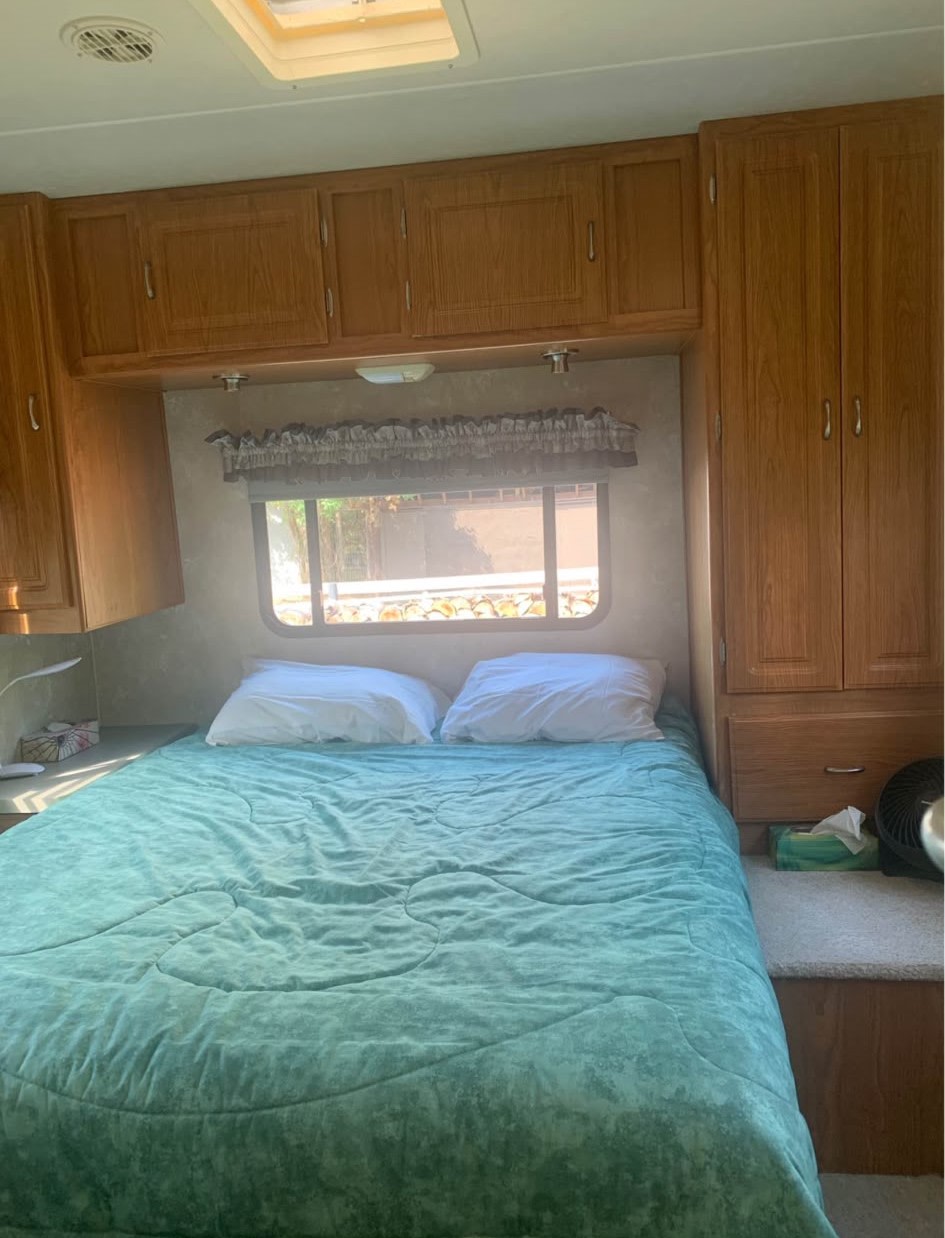 Cozy RV rental bedroom featuring a teal comforter, white pillows, overhead cabinets, and a window. Ideal for camper travel.. Coachmen Freelander Deluxe/Premier 2004