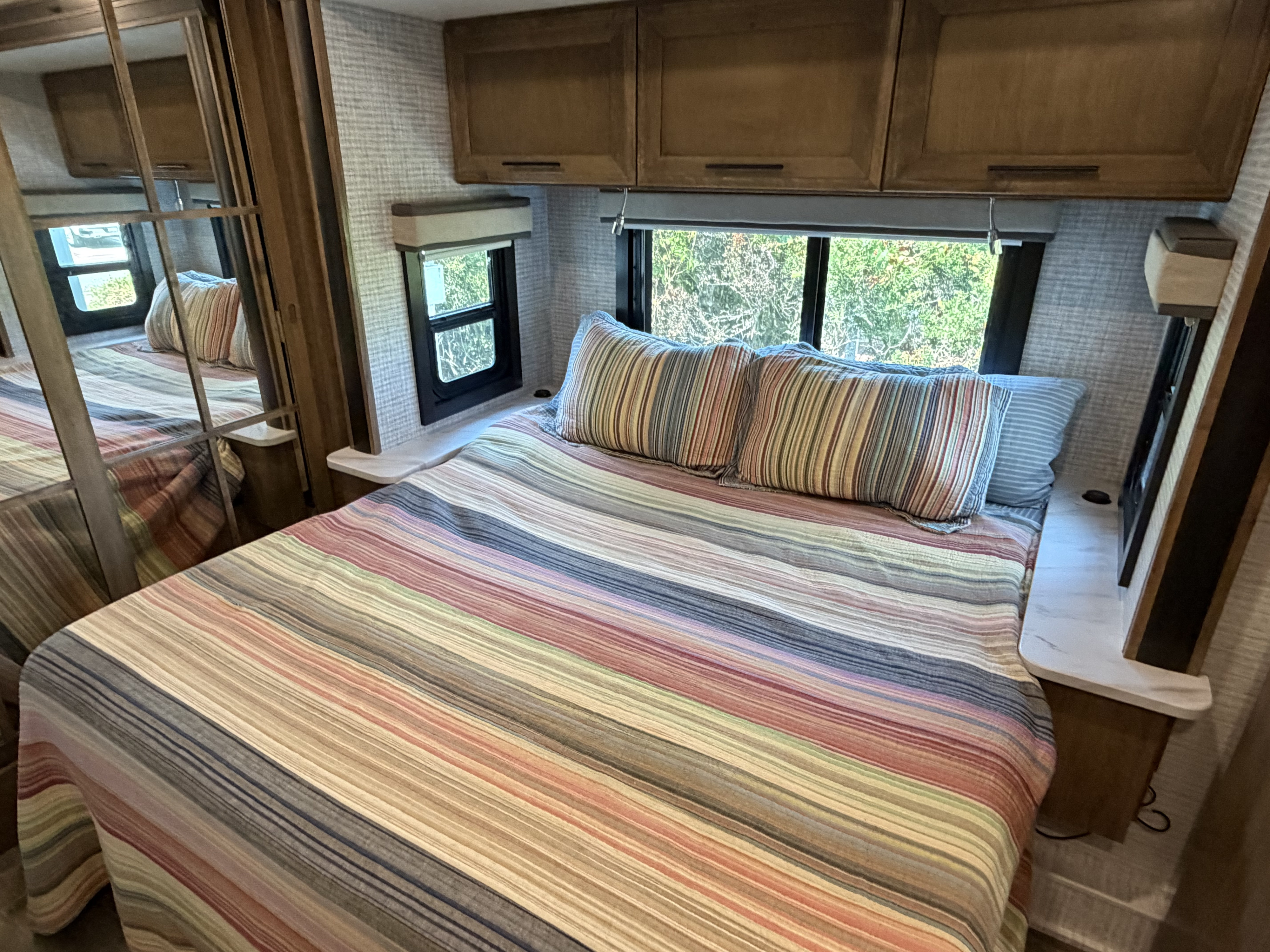 Cozy RV rental bedroom featuring a striped bedspread, pillows, overhead cabinets, and windows with green views.. Entegra Coach Esteem XL 2024