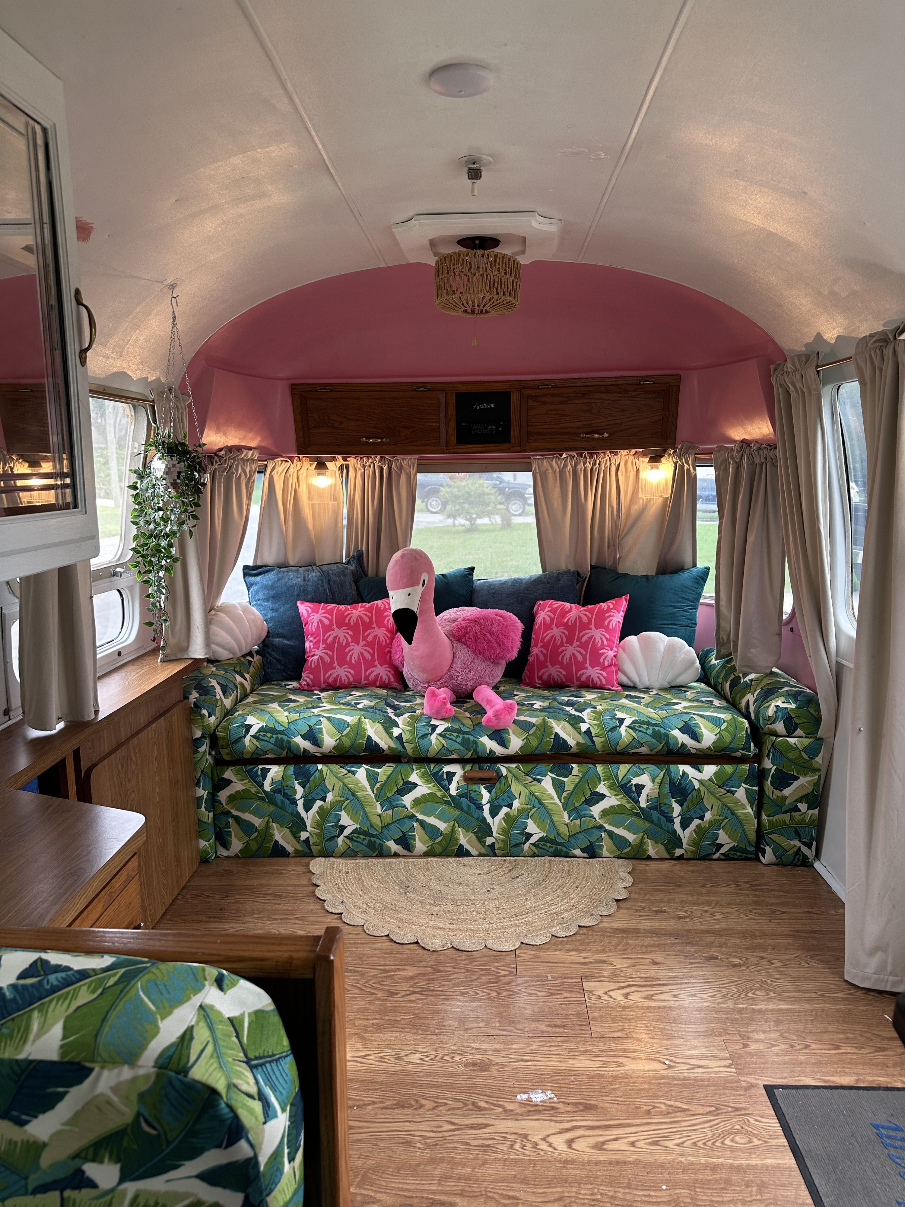 Bright rv rental interior: pink ceiling, tropical leaf sofa, flamingo, and wood floor. Cozy, colorful glamping space.. Airstream Excella 1991