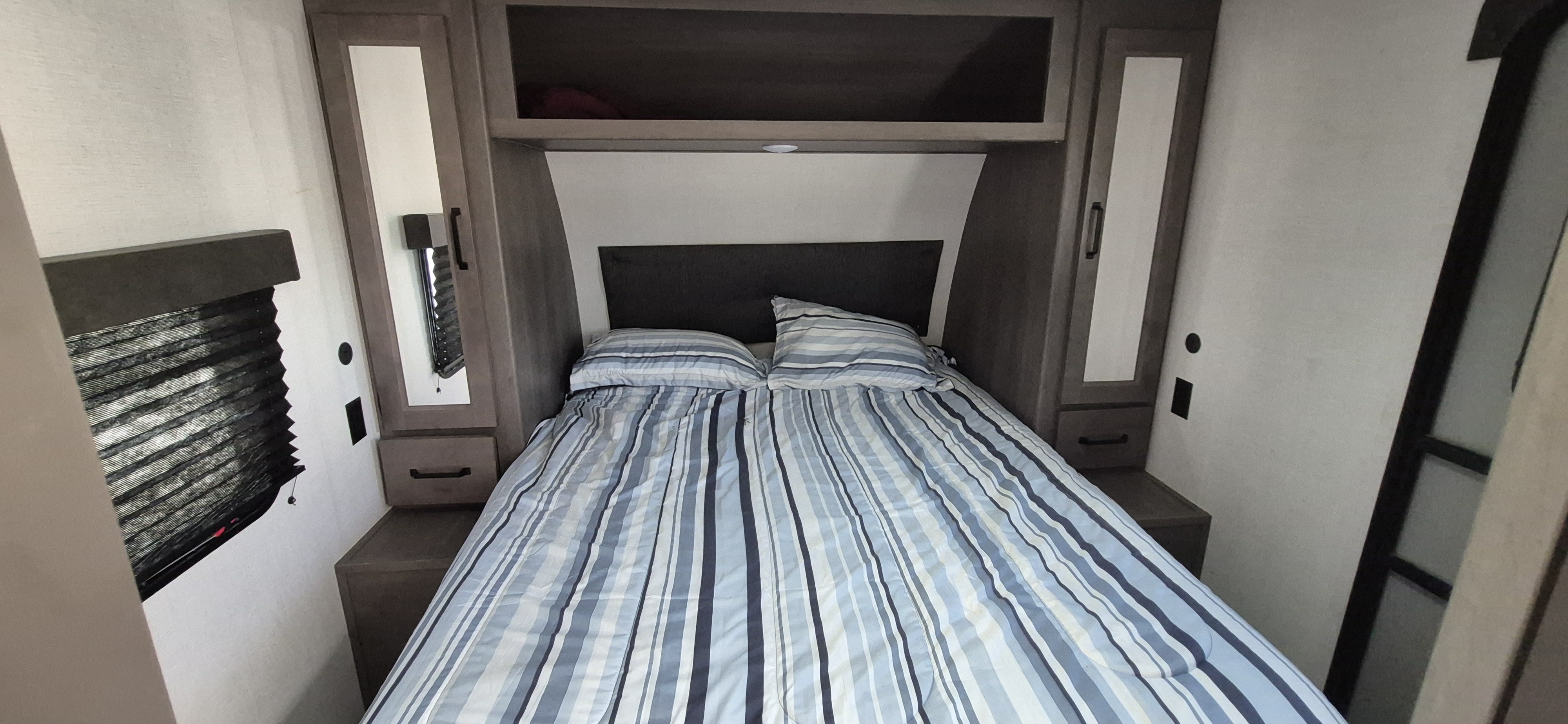 This rv rental bedroom features a comfy bed with striped bedding, overhead storage, and a window.. Grand Design Transcend Xplor 2024