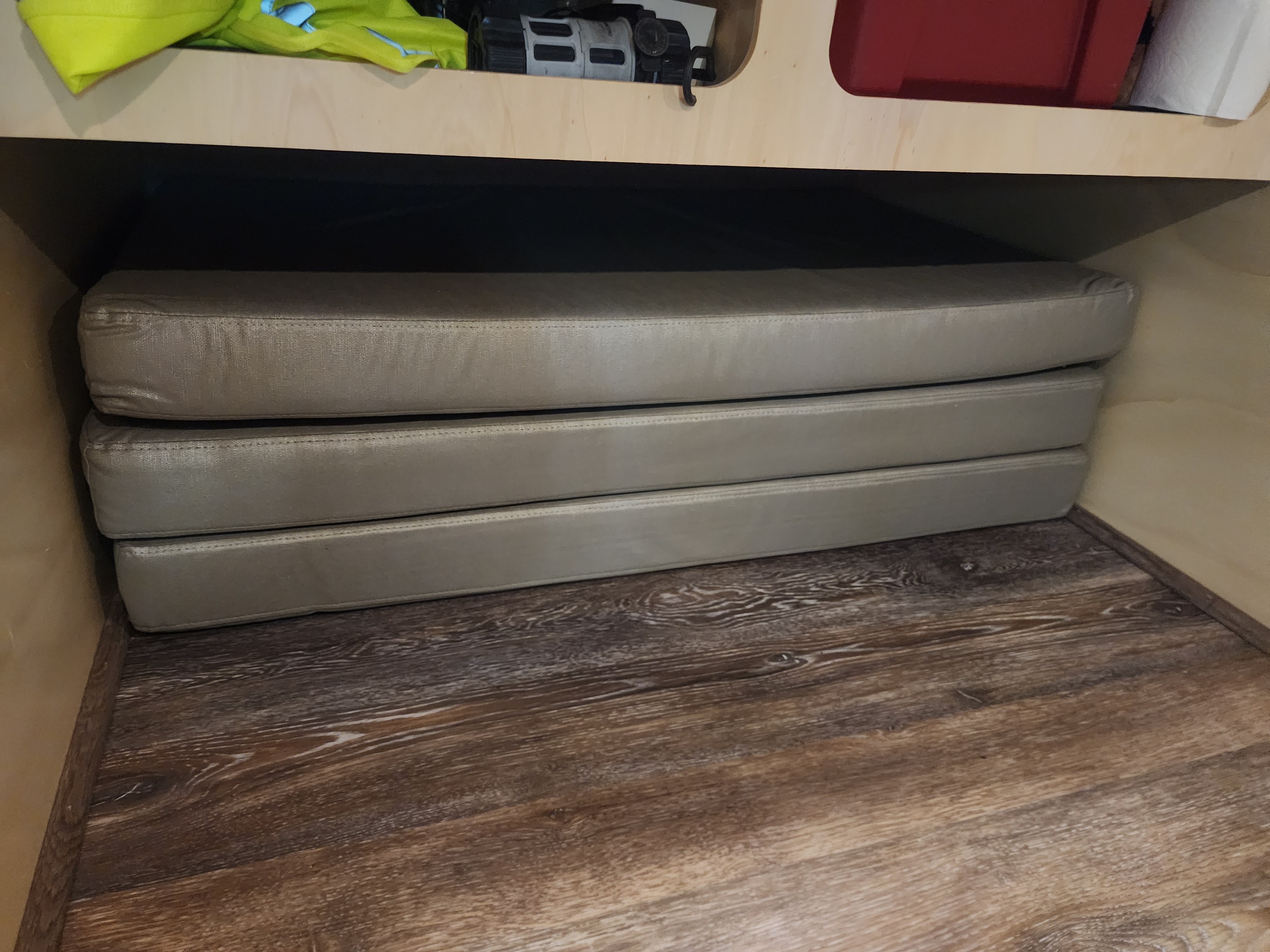Stacked beige cushions in an RV rental storage area under a wooden shelf, providing versatile seating or sleeping options.. Off Grid Trailers Off Grid Trailers Trailer 2021