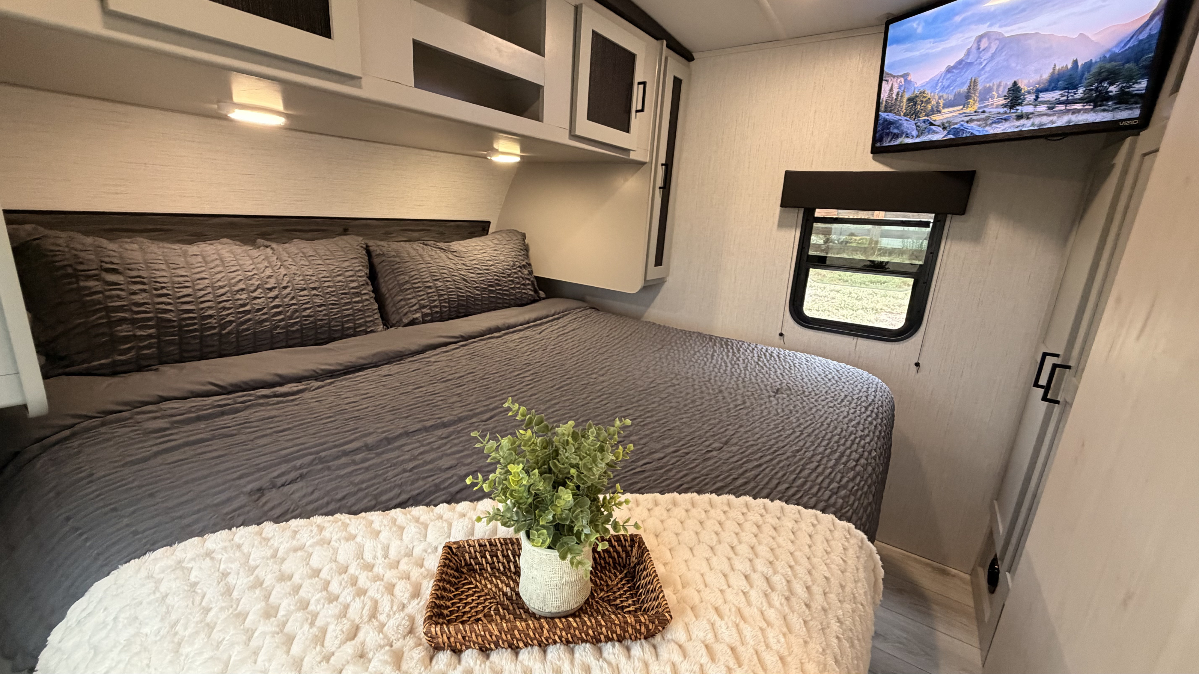 A comfortable RV rental bedroom features a gray bed with textured bedding, overhead cabinets, a TV, and a window view.. Heartland RVs North Trail 2022