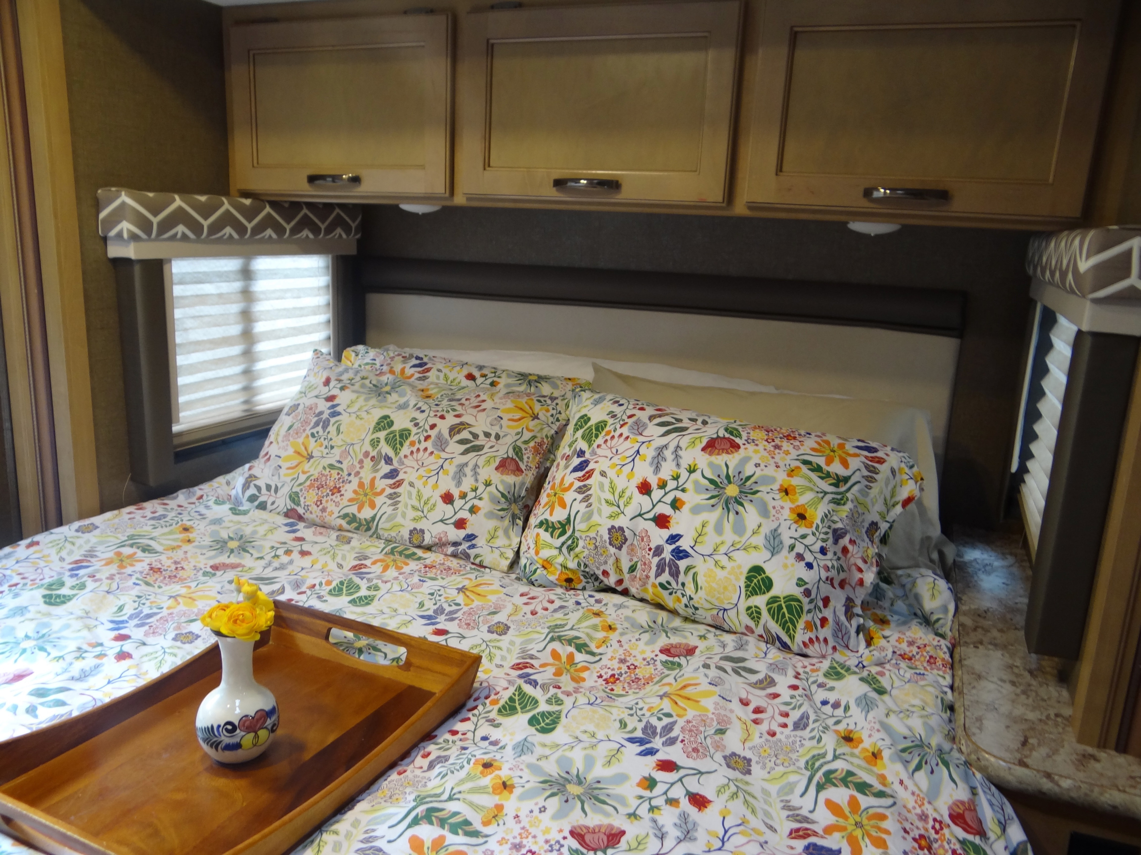 Cozy RV rental bedroom with colorful floral bedding, overhead cabinets, and windows with blinds, featuring a decorative tray on the bed.. Thor Daybreak 2019