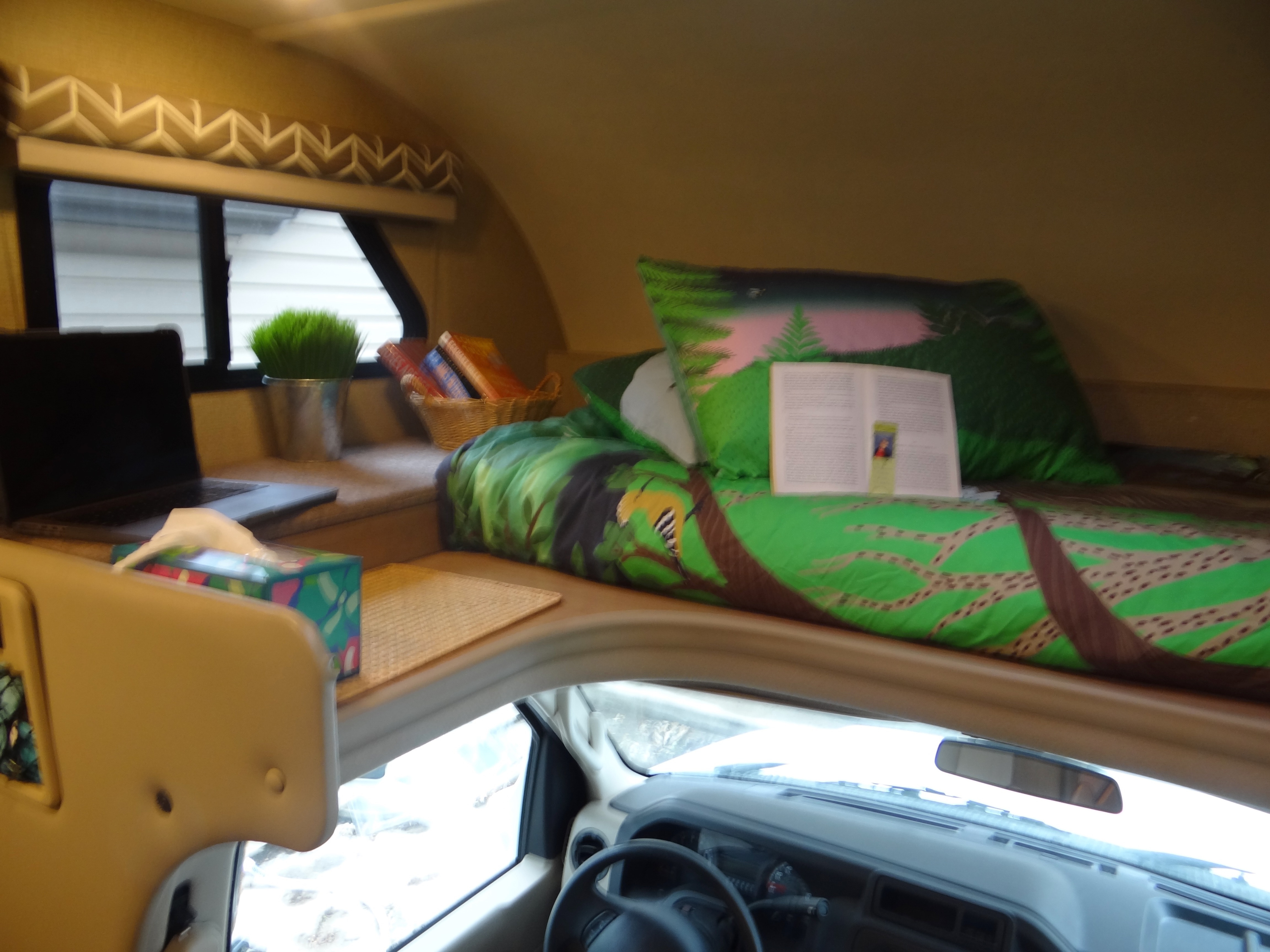 Cozy RV rental interior: overhead bunk bed with an open book, laptop, and essential travel items for comfortable journeys.. Thor Daybreak 2019