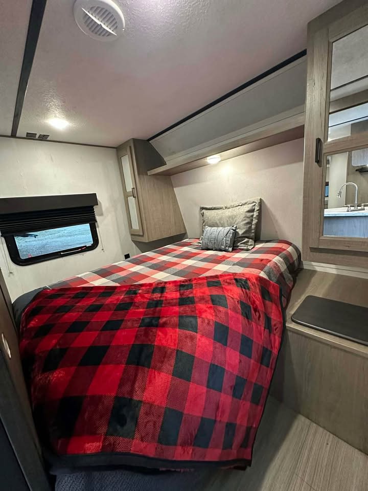 Charming RV rental bedroom with a cozy red plaid blanket, window, overhead storage, and mirrored closet.. Keystone RV Hideout 2021