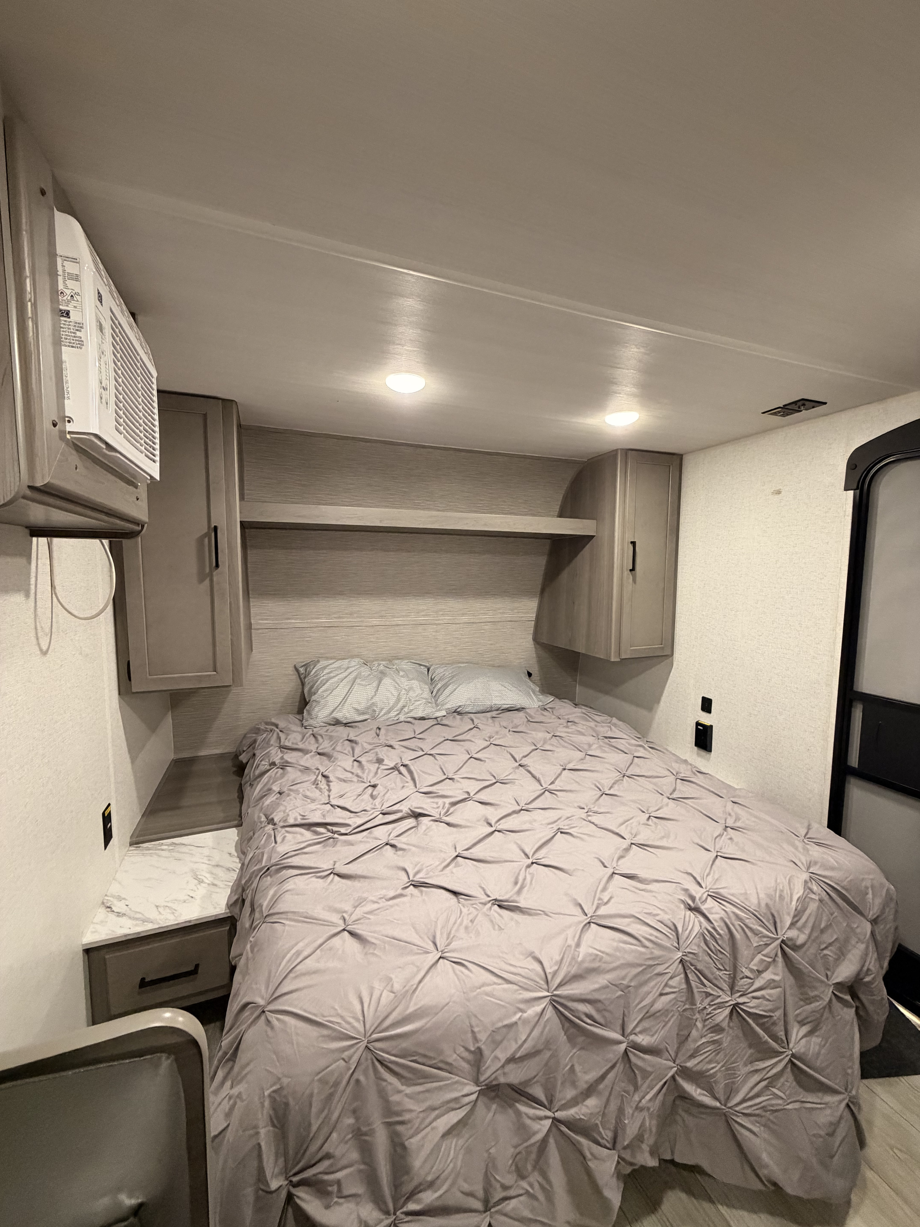 Cozy RV rental bedroom showing a bed with a grey comforter, overhead cabinets, a nightstand, and a wall-mounted AC unit.. Keystone RV Hideout Sport 2025