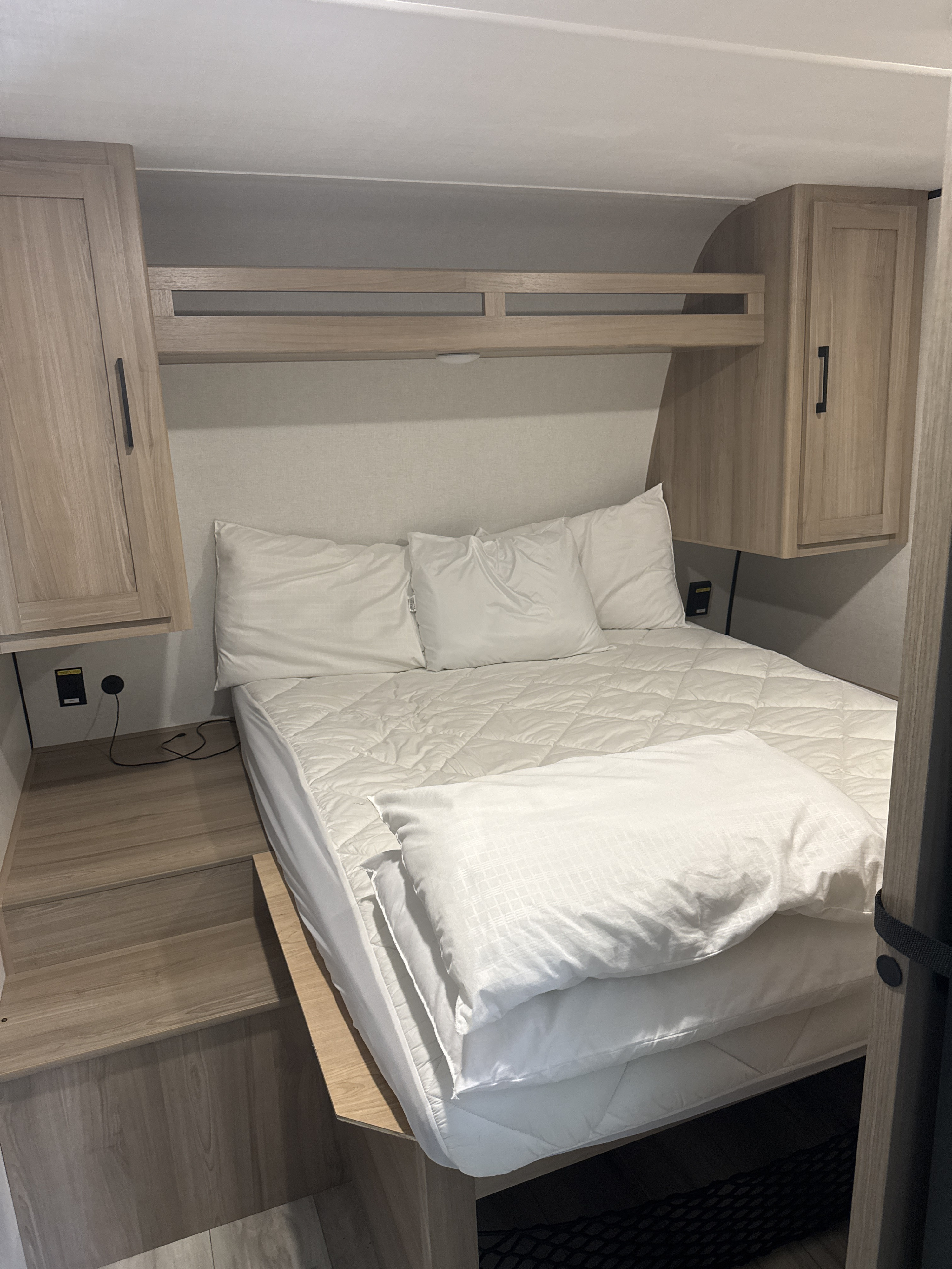 Cozy RV rental bedroom with a compact bed, white bedding, overhead wood cabinets, and built-in steps for easy access.. Keystone RV Coleman Light 2026