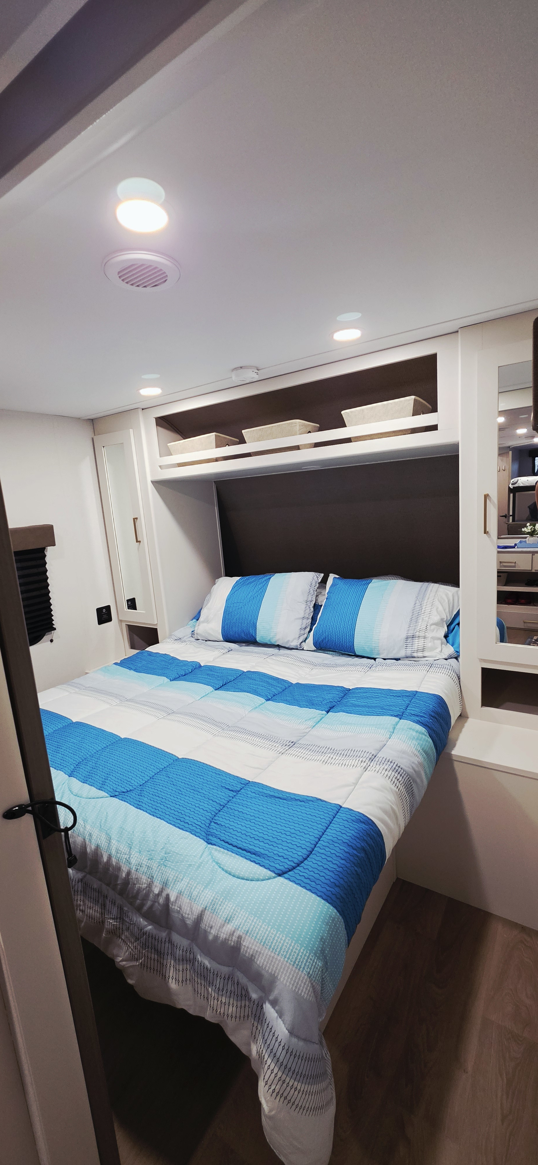Bright RV rental bedroom with blue striped bed, built-in storage, mirrors, and ceiling lights for comfortable travel.. Grand Design Transcend Xplor 26BHX 2027
