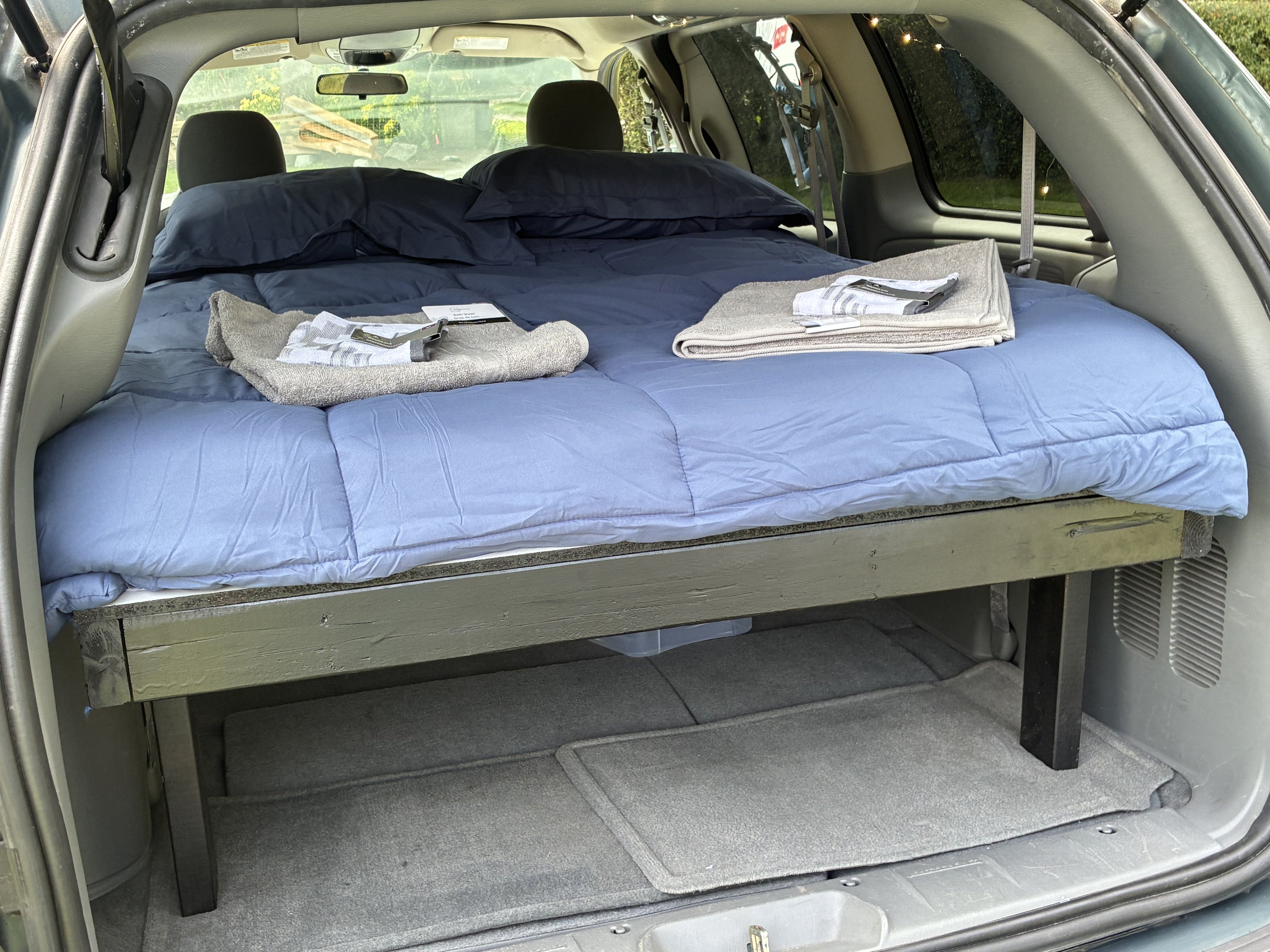 Comfy bed setup in a minivan campervan, ideal for an RV rental or car camping. Features a mattress, blue bedding & towels.. Dodge Caravan 2006