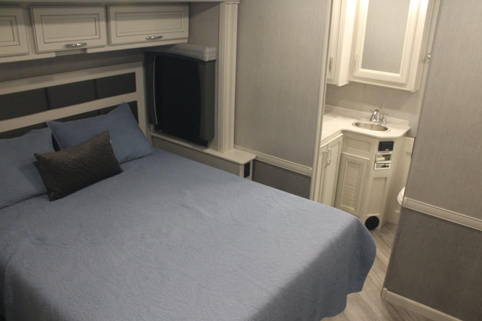 Compact RV rental interior: bedroom with blue bedding, built-in TV, and a functional bathroom vanity. Ideal for travel.. Holiday Rambler Vacationer 2023