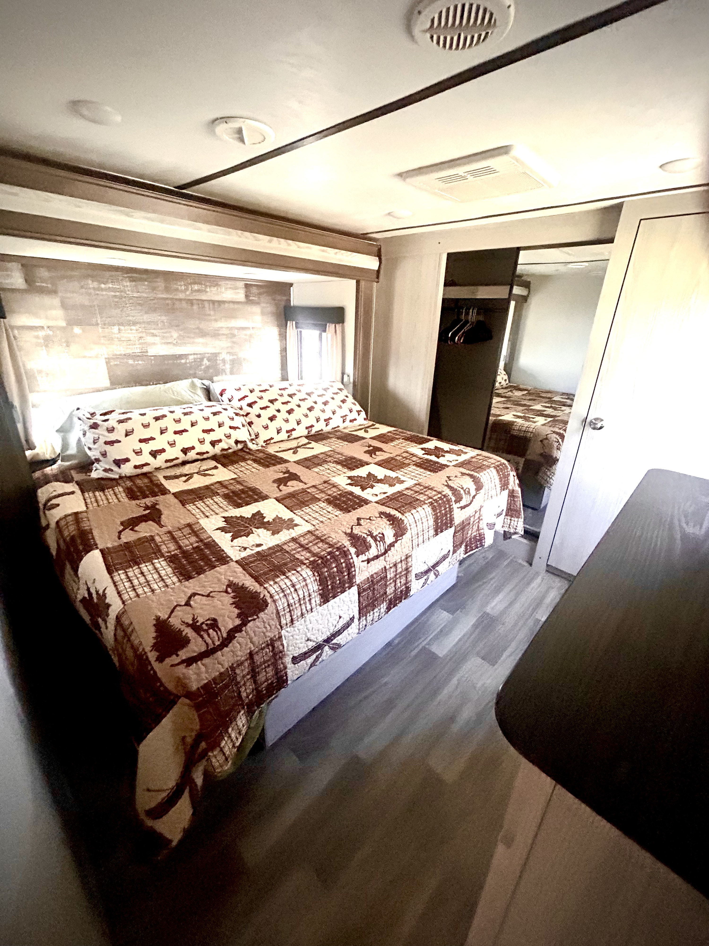 Cozy RV rental bedroom features a rustic quilt with deer & maple leaf patterns, wood headboard, and a mirrored closet.. Keystone RV Montana High Country 2020