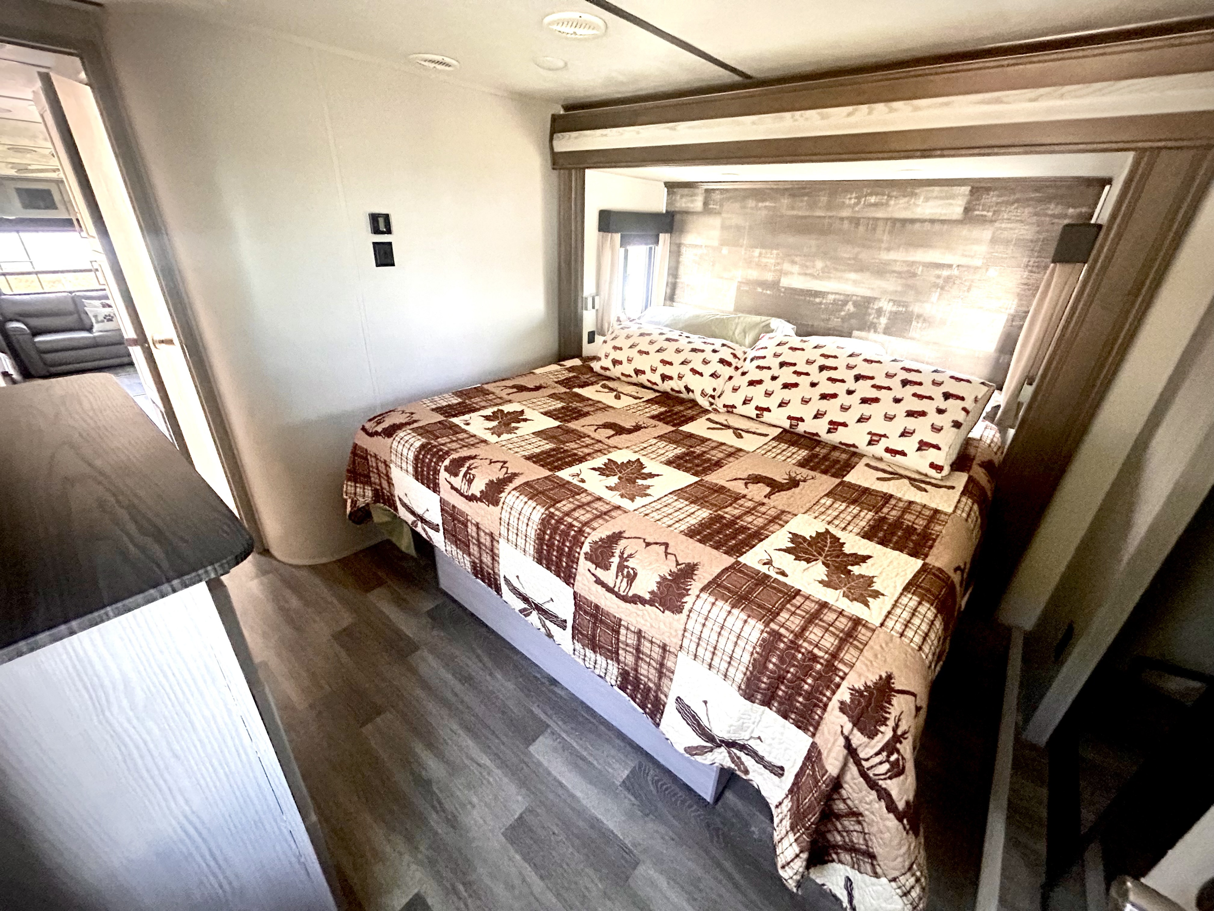 View of an RV rental bedroom with a patterned quilt, dark wood headboard, and a glimpse into the living area.. Keystone RV Montana High Country 2020