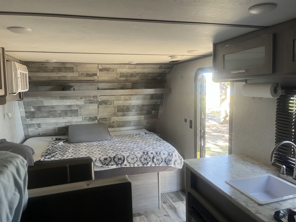 Inside an RV rental, featuring a comfortable bed, an upper bunk, a compact kitchen with sink, and a bright view through the open door.. Dutchmen Coleman Lantern LT 2022