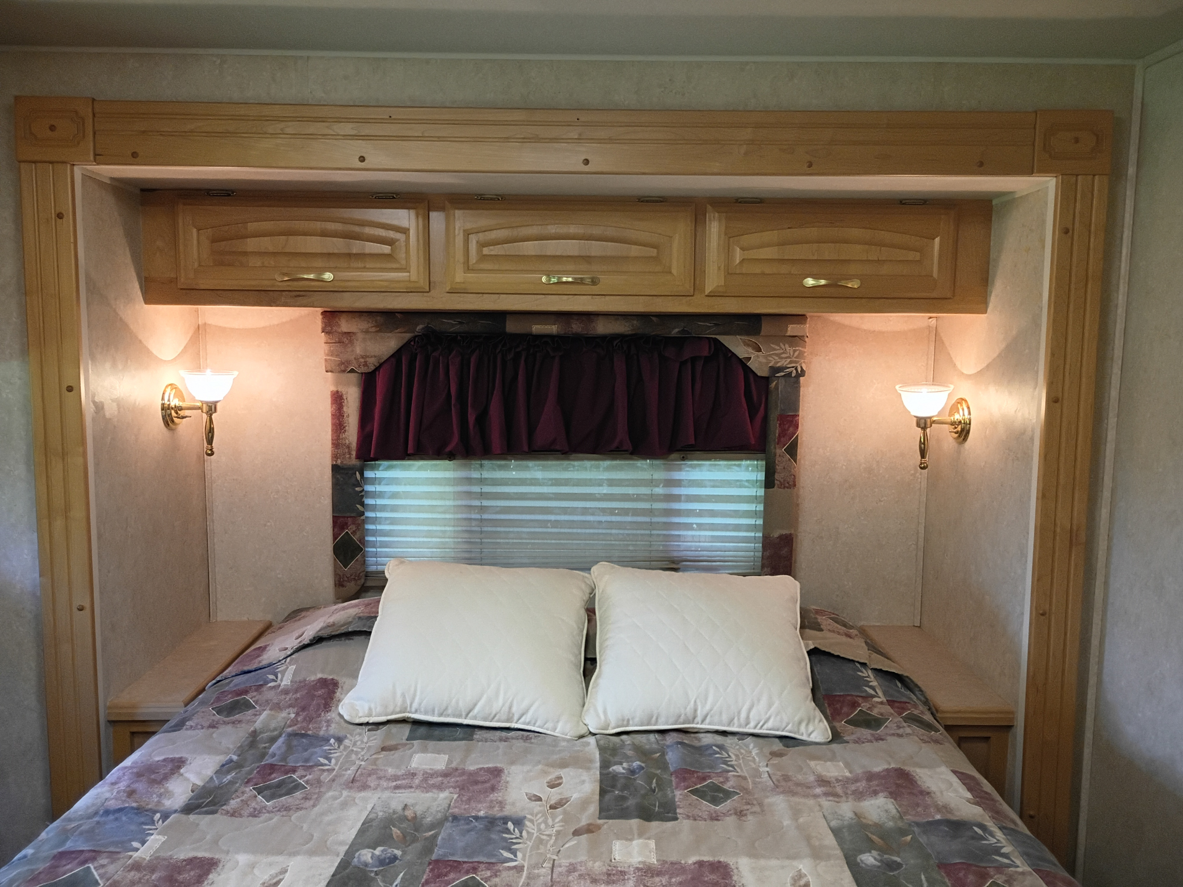 Comfortable RV rental bedroom featuring a bed with patterned comforter & pillows, overhead wood cabinets, window with blinds, and warm wall sconces.. Georgie Boy Cruise-Master 2003