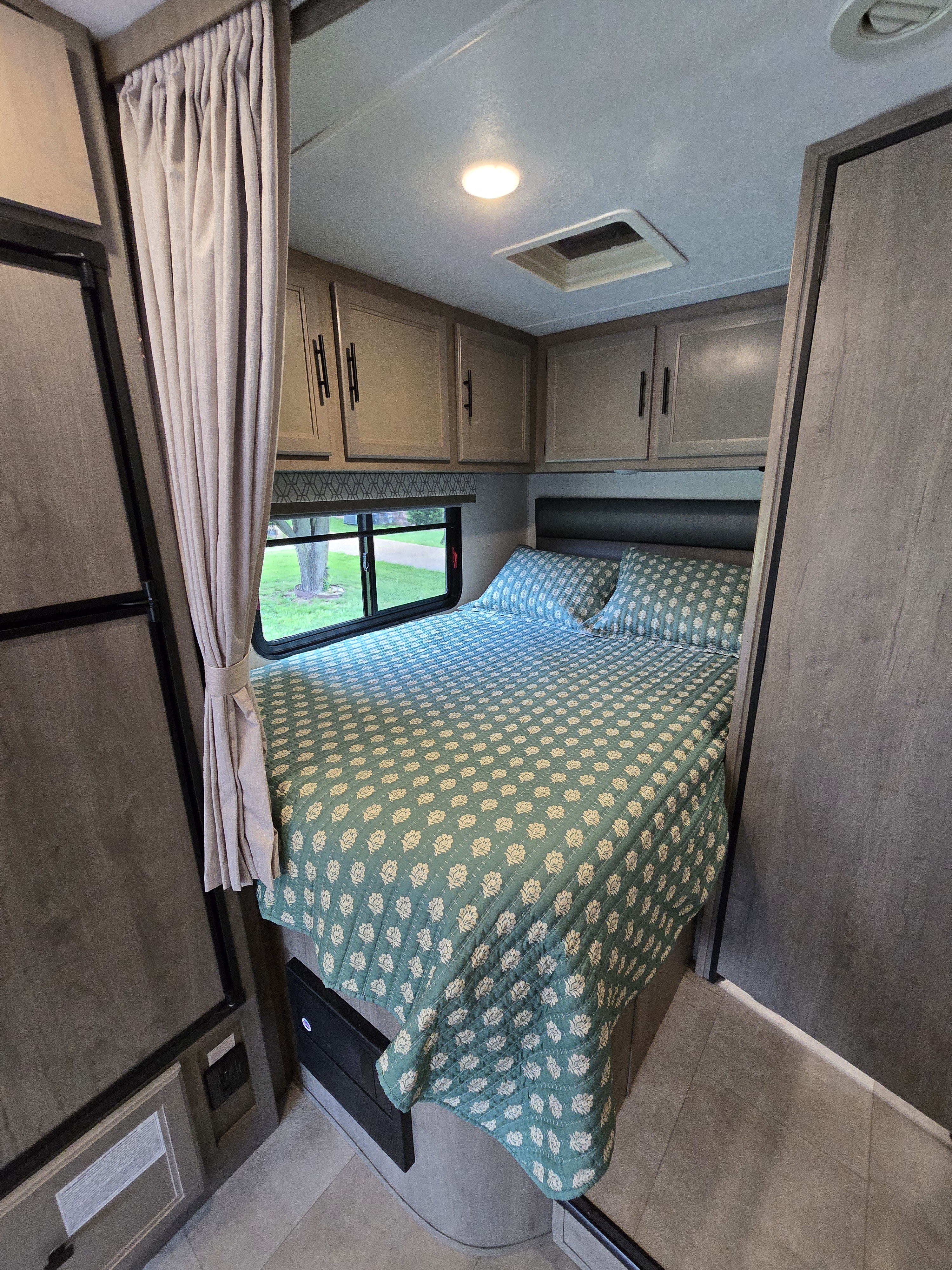 Cozy RV rental bedroom featuring a teal patterned bed, overhead cabinets, a window with outdoor view, and a privacy curtain.. Forest River Coachmen Prism 2020