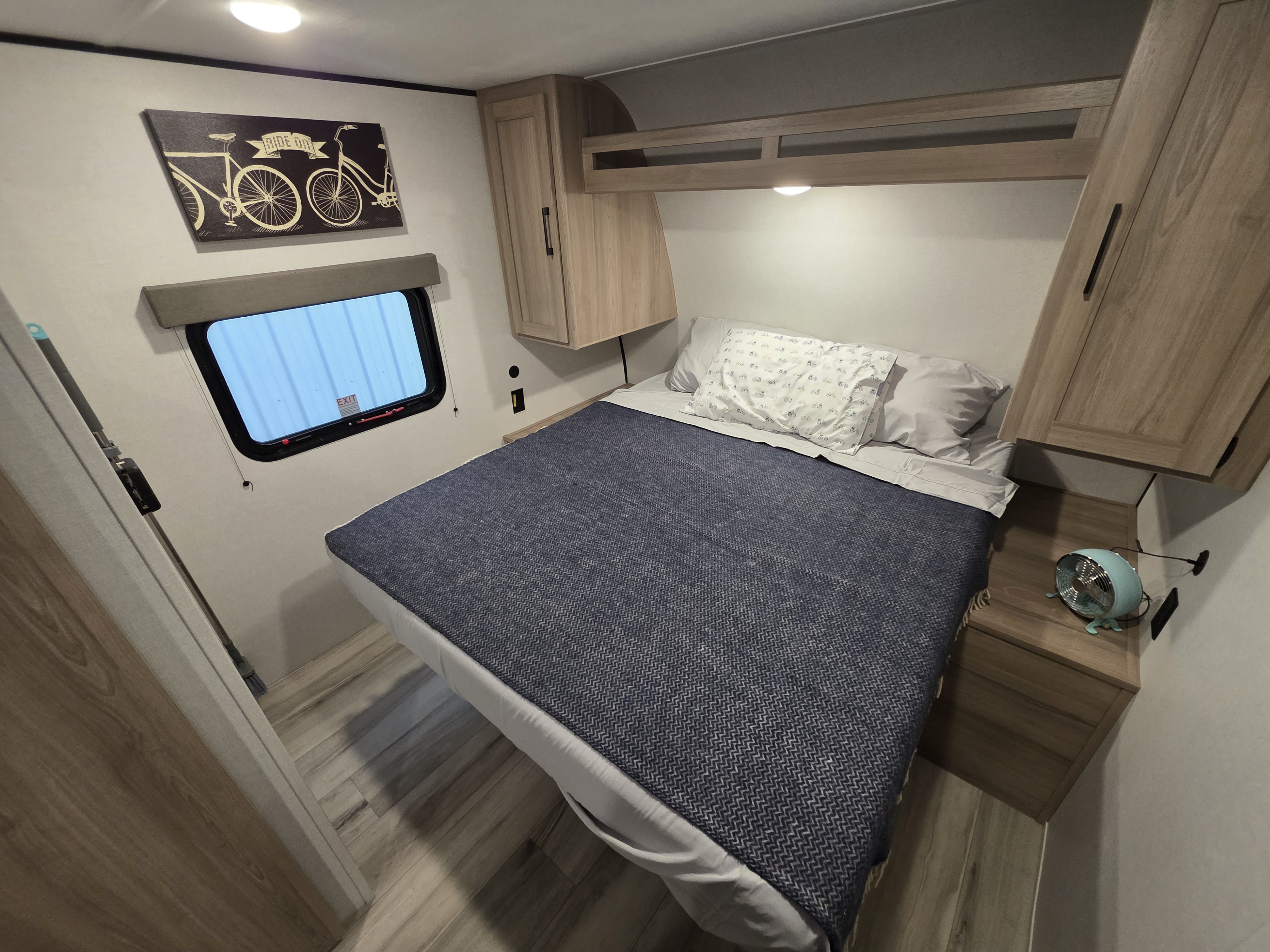 This RV rental bedroom offers a comfortable bed, overhead cabinets, a window, and charming bicycle wall art.. Keystone RV Coleman 2026
