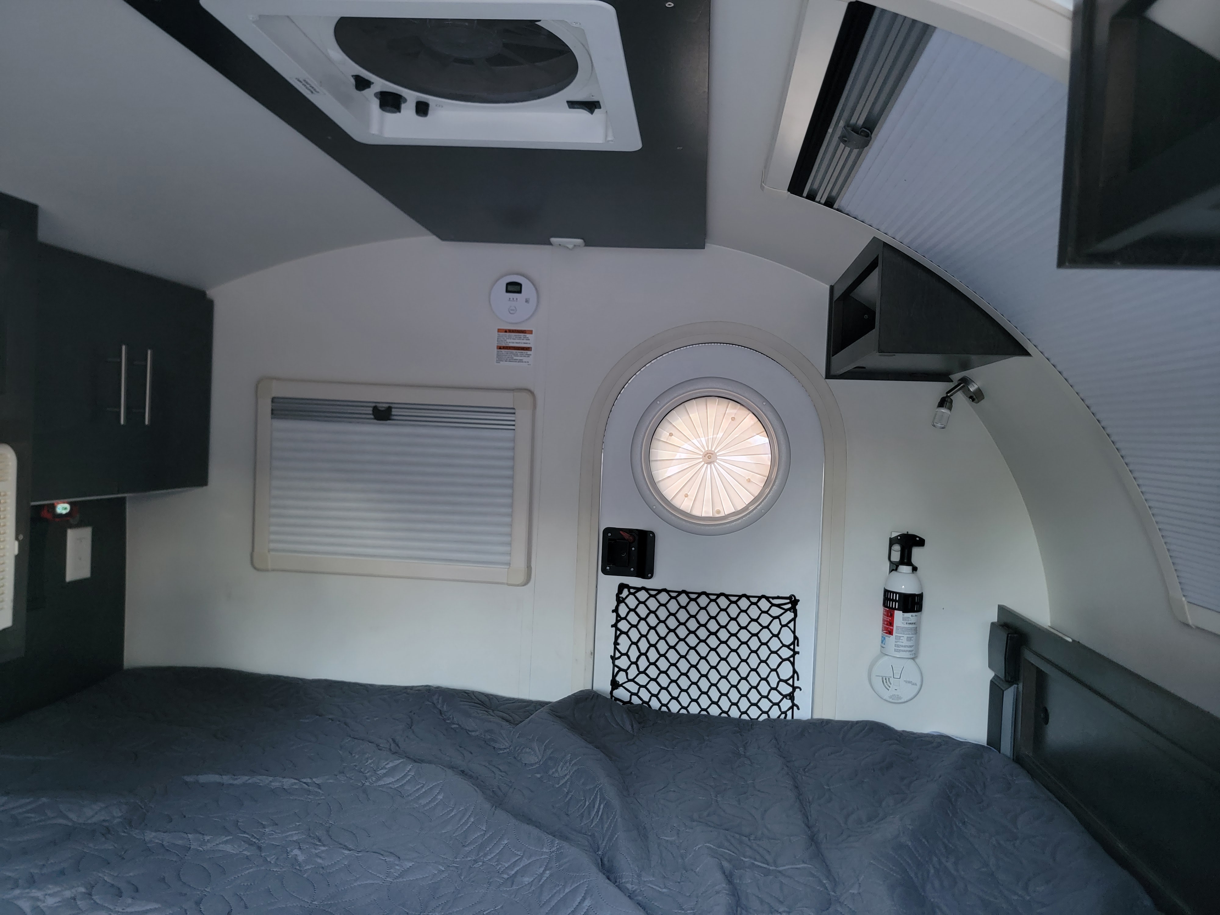 Cozy RV rental interior with bed, window, round door, overhead fan, carbon monoxide detector, and fire extinguisher visible.. nuCamp T@G 2021