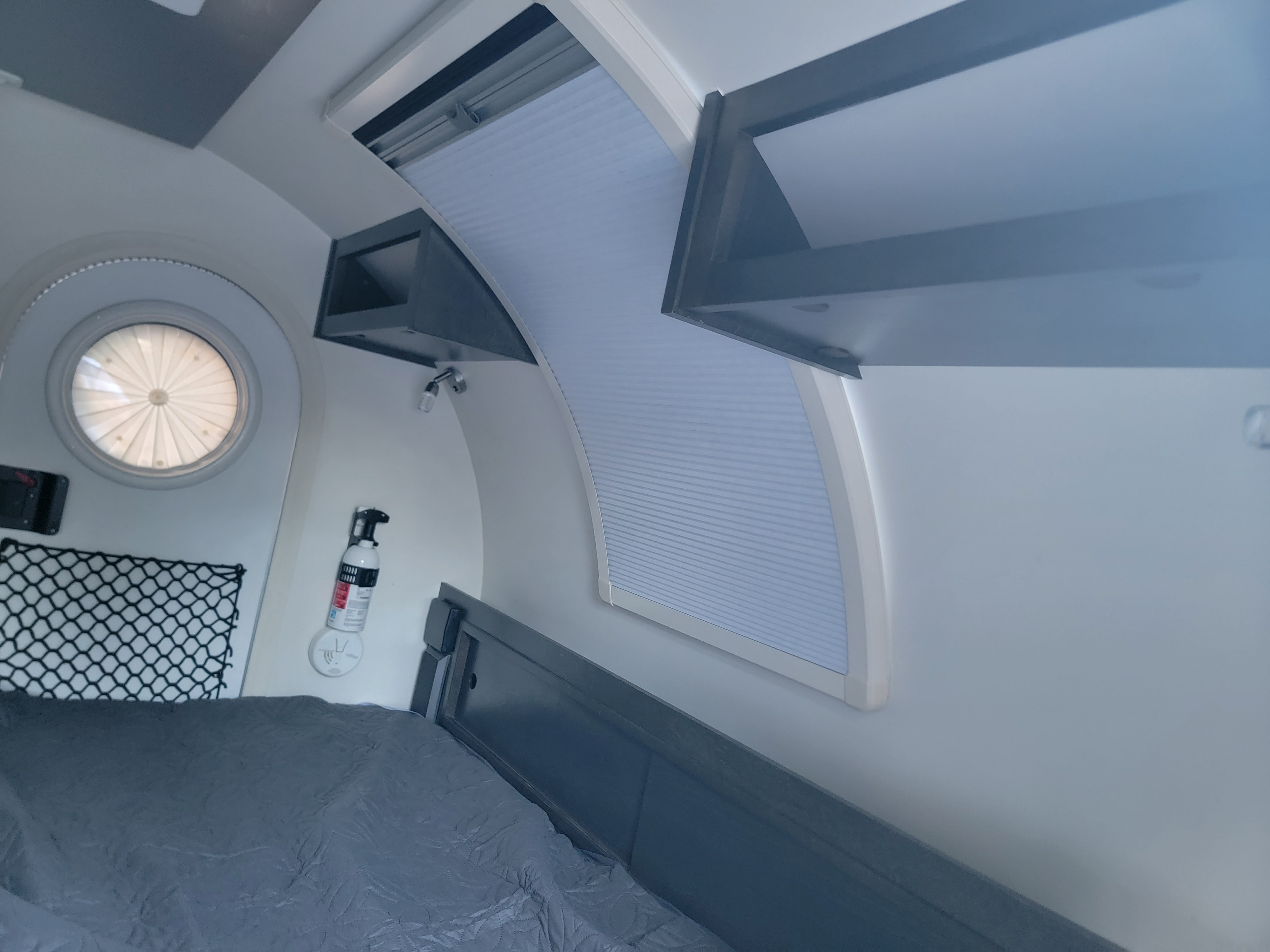 Cozy RV rental interior with bed, round window, pleated shades, overhead storage, fire extinguisher, and smoke detector.. nuCamp T@G 2021