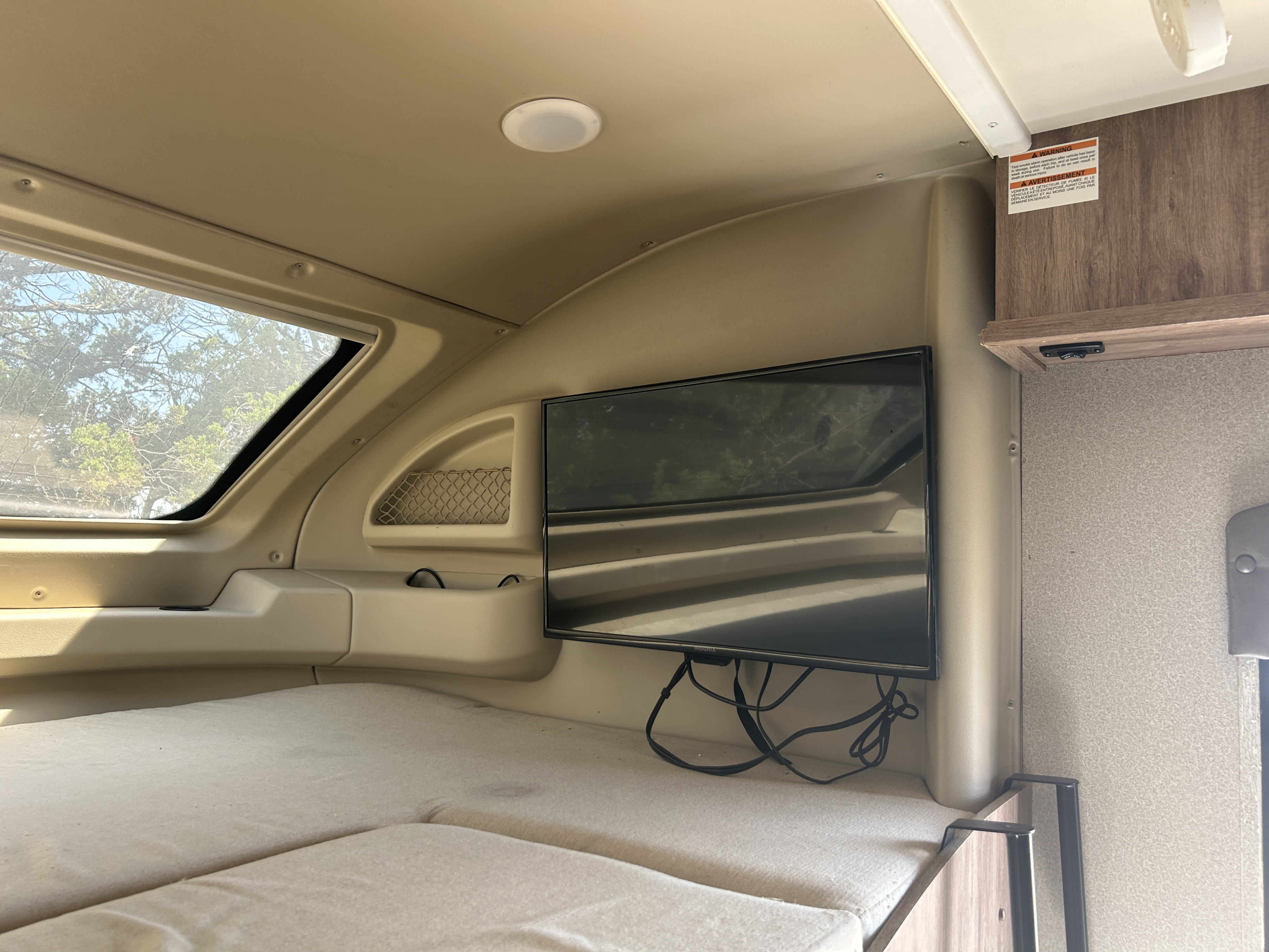 Cozy RV rental sleeping area with window views, mounted TV, and a safety warning sticker above storage.. Entegra Coach Odyssey 2019