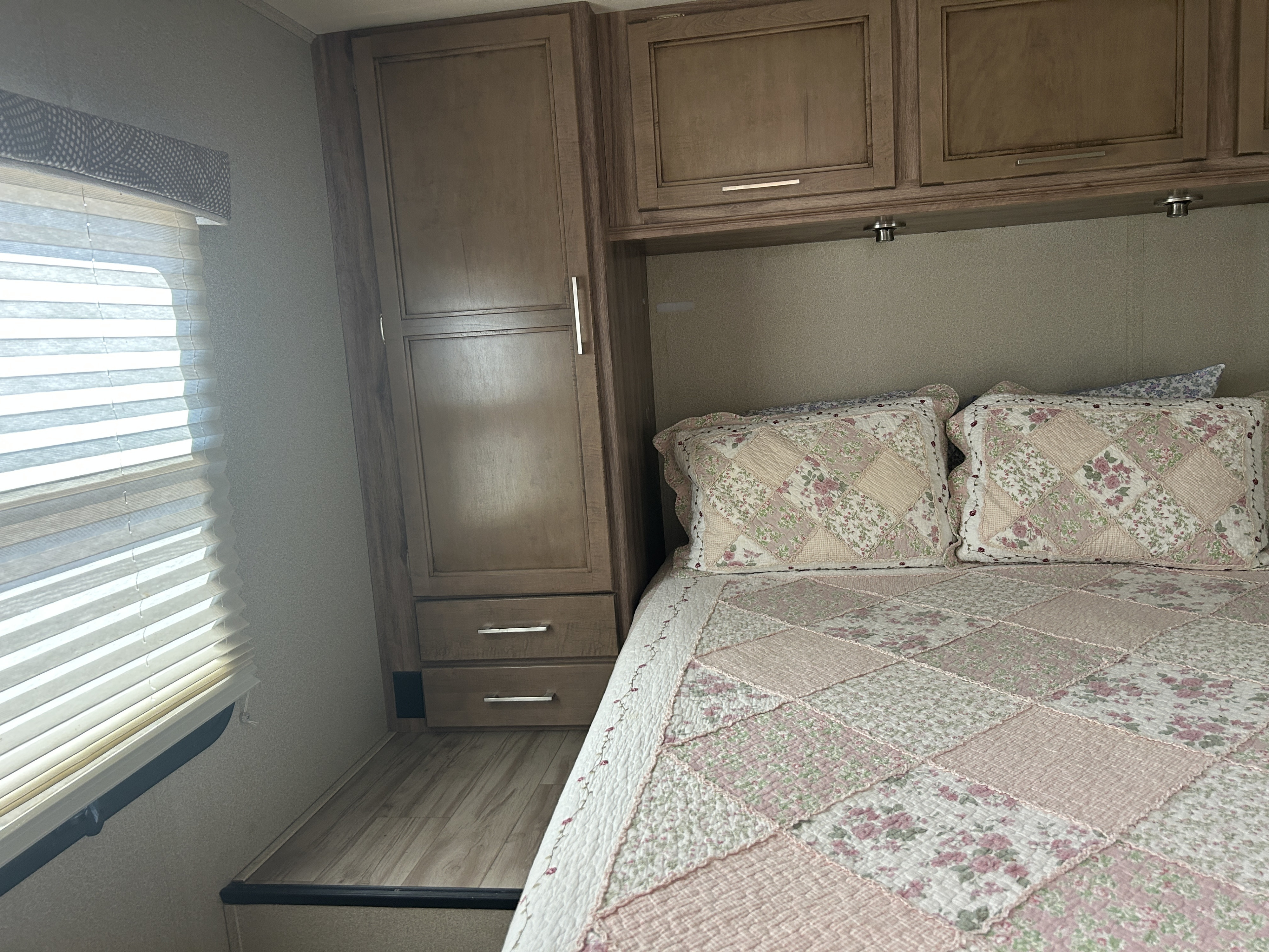 Cozy RV rental bedroom featuring a patchwork quilt and pillows, a window with blinds, and wooden cabinets.. Entegra Coach Odyssey 2019