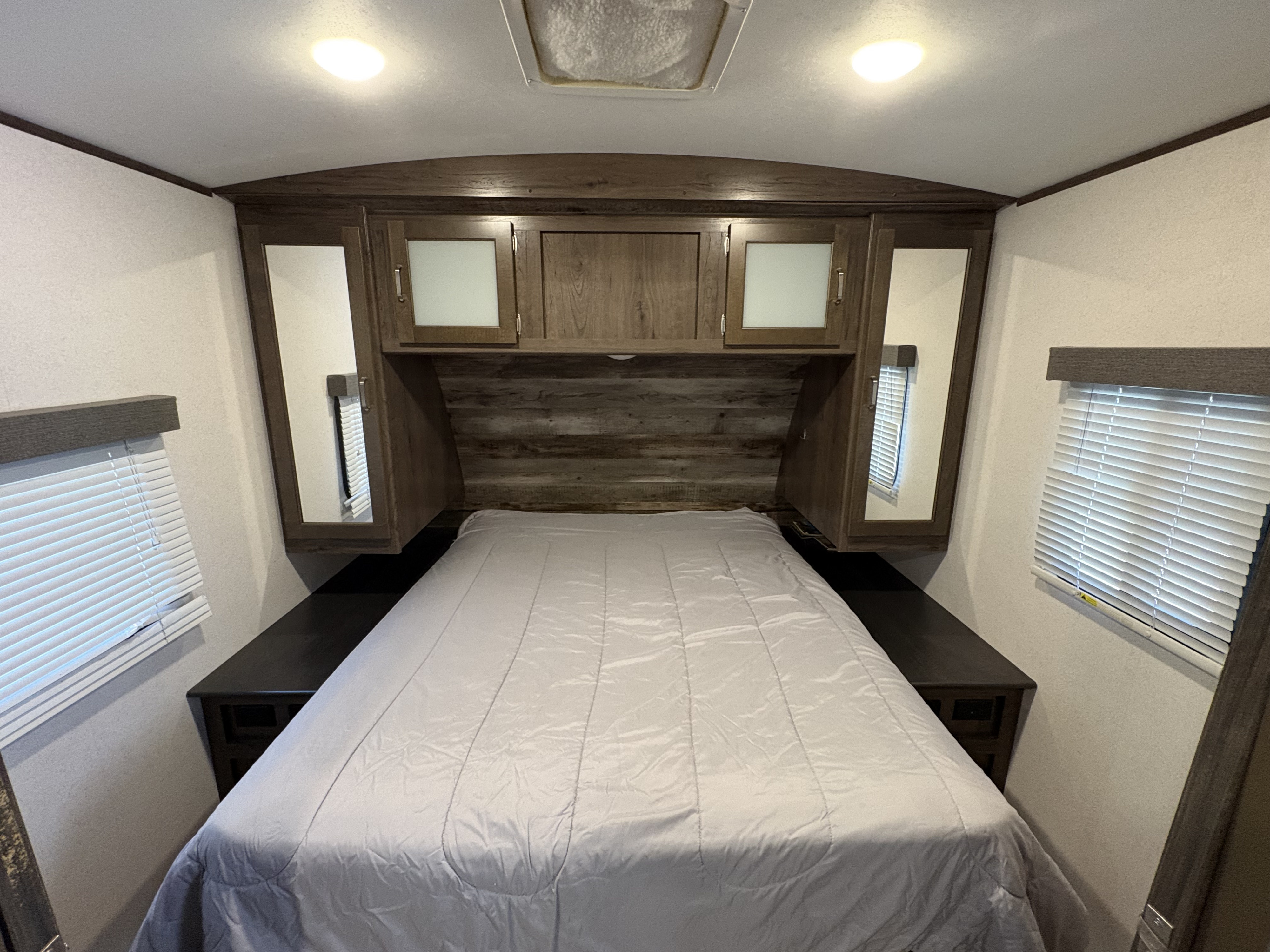 Inside a comfortable RV rental bedroom featuring a full bed, overhead storage cabinets, and side windows with blinds.. Keystone RV Passport Grand Touring 2020