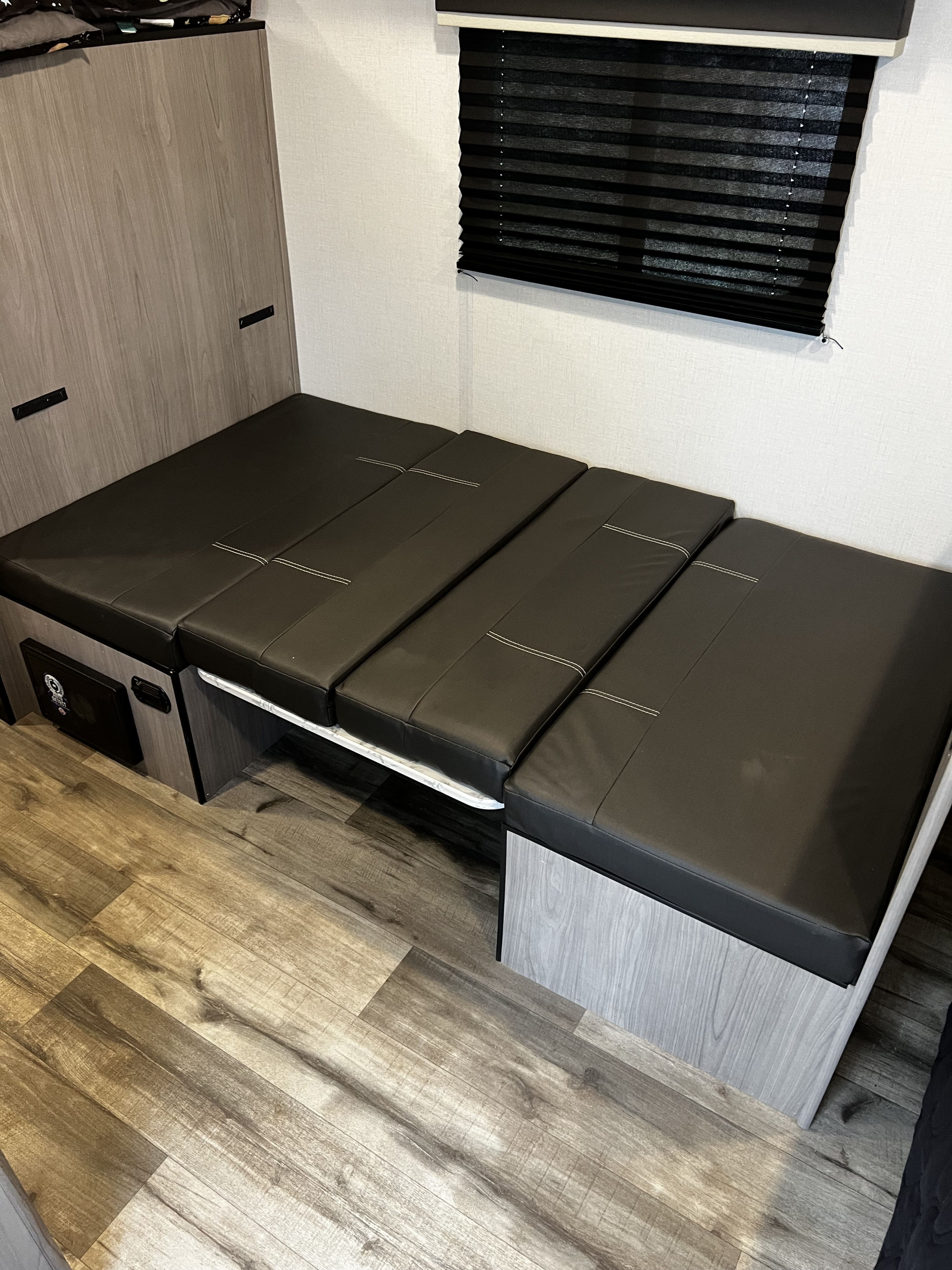 RV rental interior featuring a dark cushioned convertible bed/seating area, wood-look floor, and window blind.. Dutchmen Coleman 2024