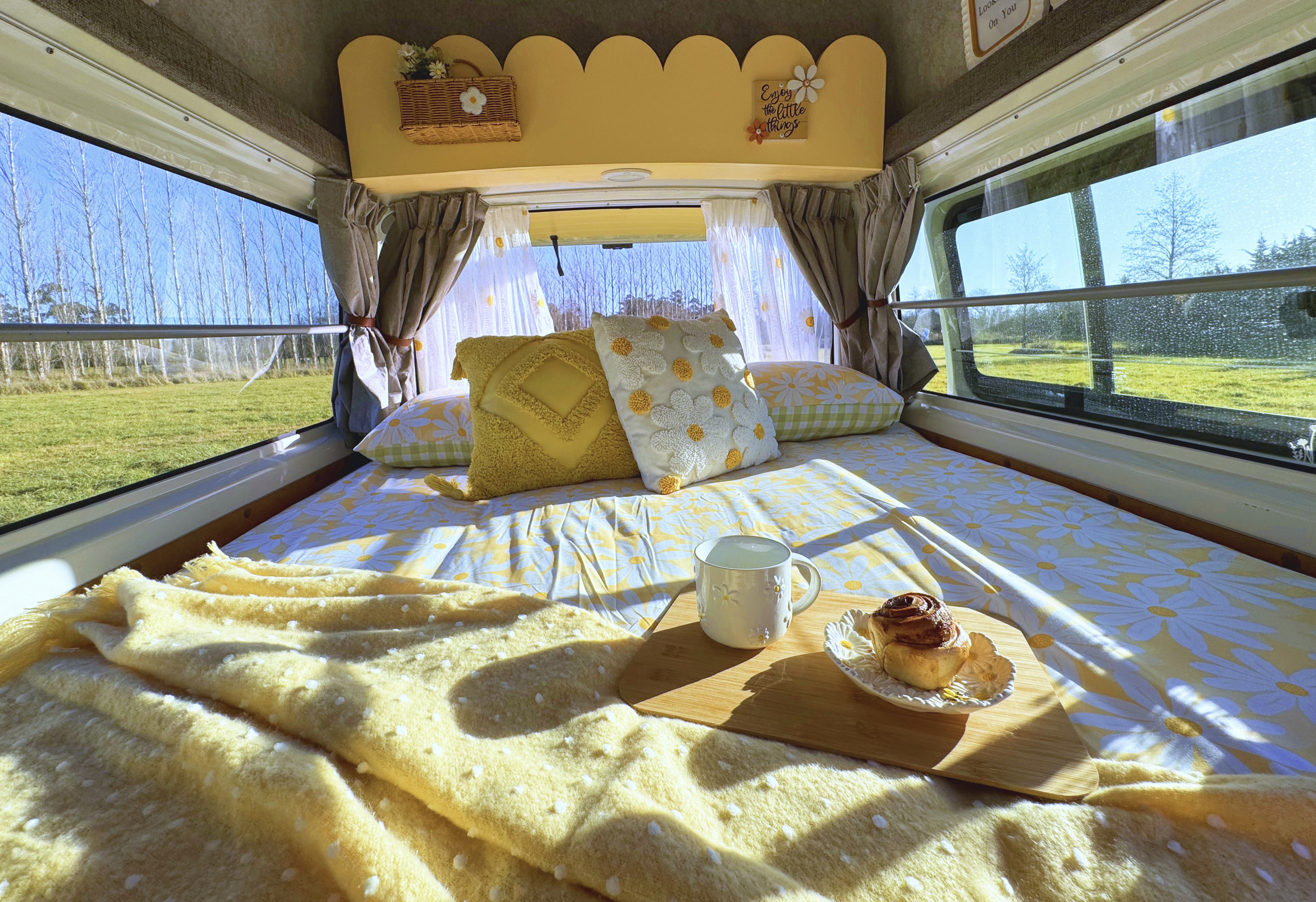 Cozy RV rental bedroom featuring bright daisy decor, breakfast in bed with a cinnamon roll, and scenic outdoor views.. Toyota Hiace 2013