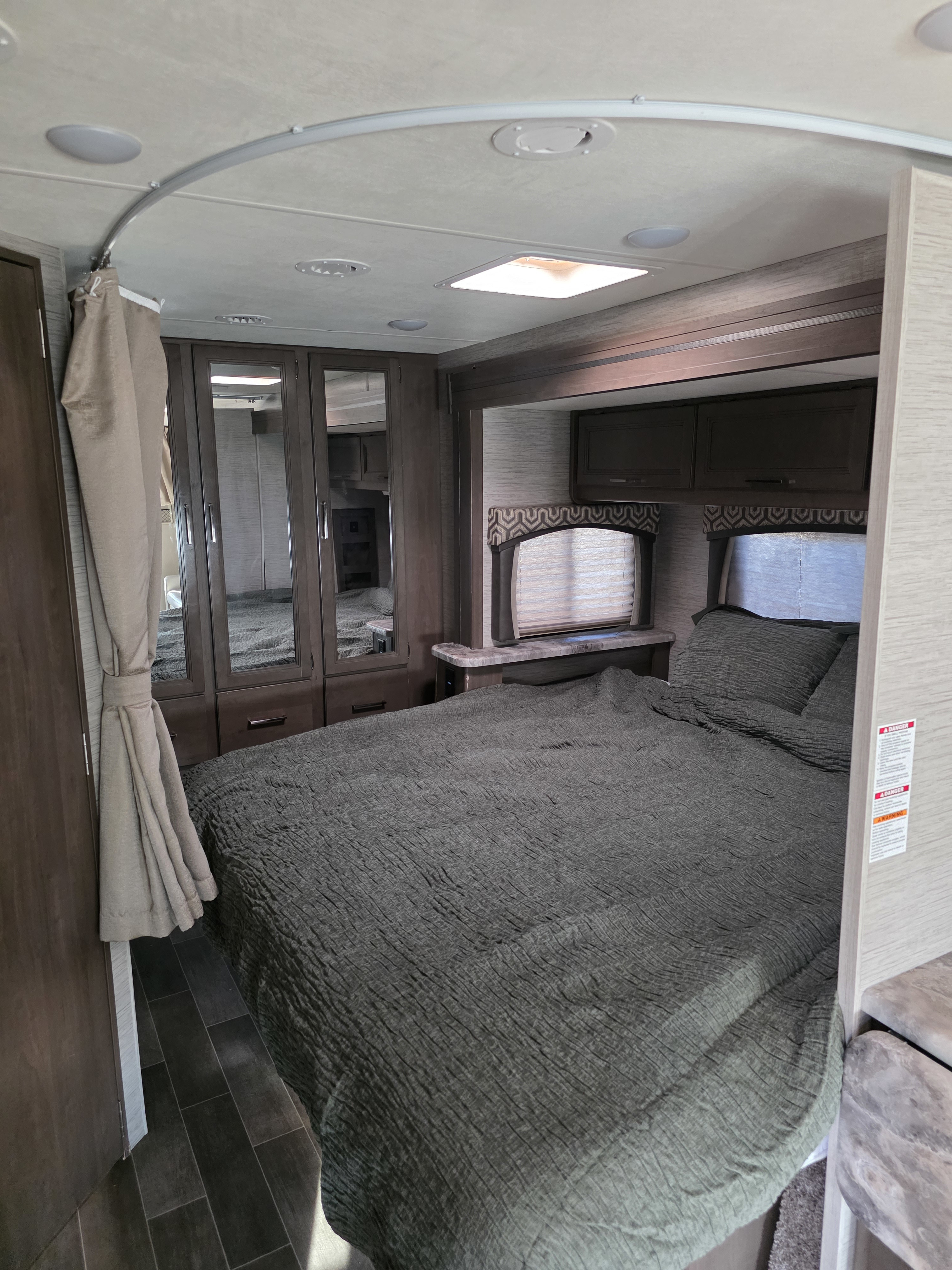 RV rental bedroom with a dark gray bed, mirrored wardrobe, overhead cabinets, and a curved ceiling track. A compact, modern space.. Thor Chateau 2021