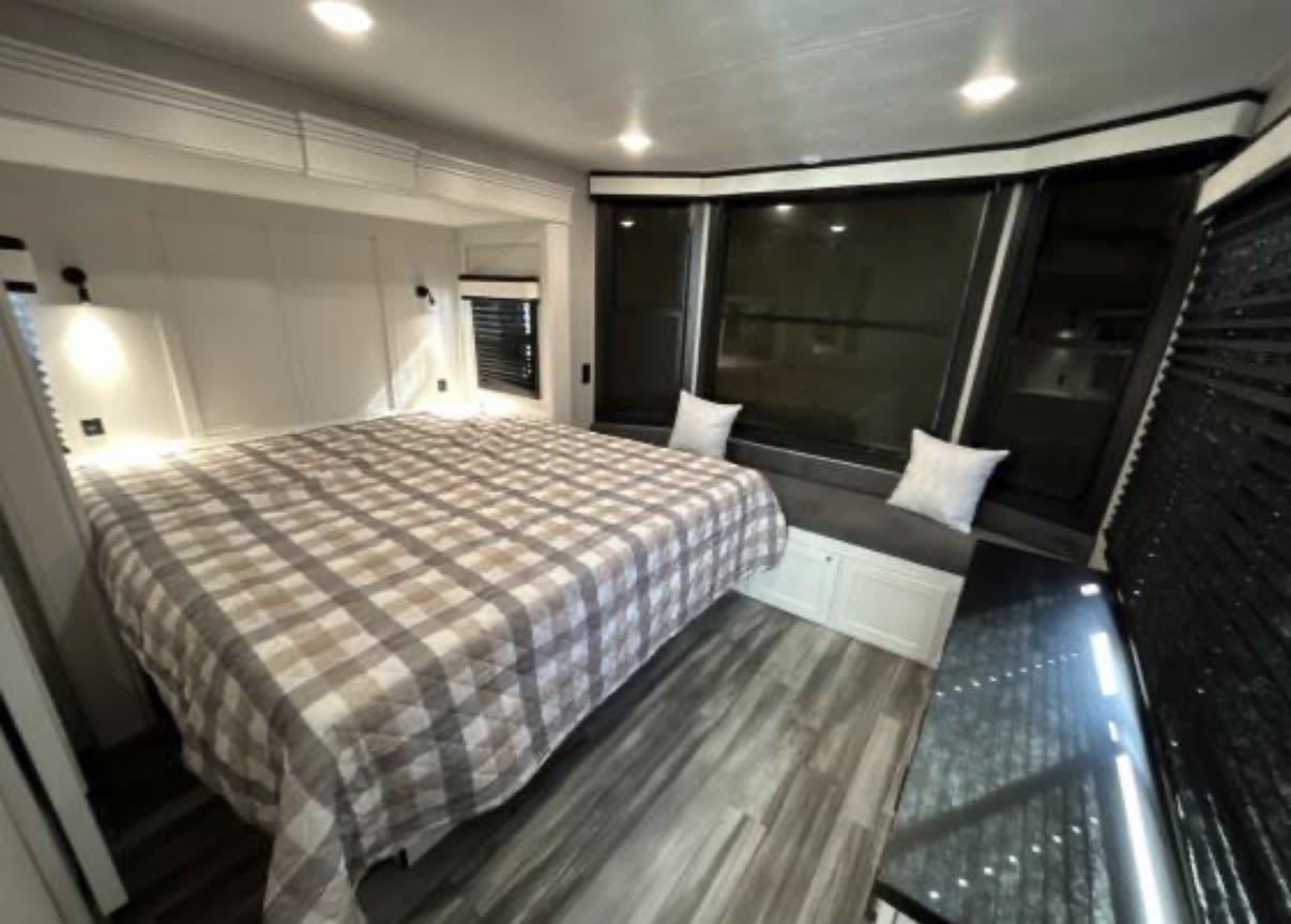 Spacious RV rental bedroom featuring a large plaid bed, window seat with pillows, windows, and wood-look flooring.. Jayco Jay Flight Bungalow 2025
