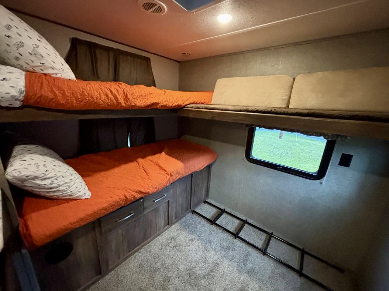 Experience comfort in this RV rental's cozy bunk beds, featuring vibrant orange bedding, a window with outdoor views, and a ladder.. Keystone RV Cougar 2018
