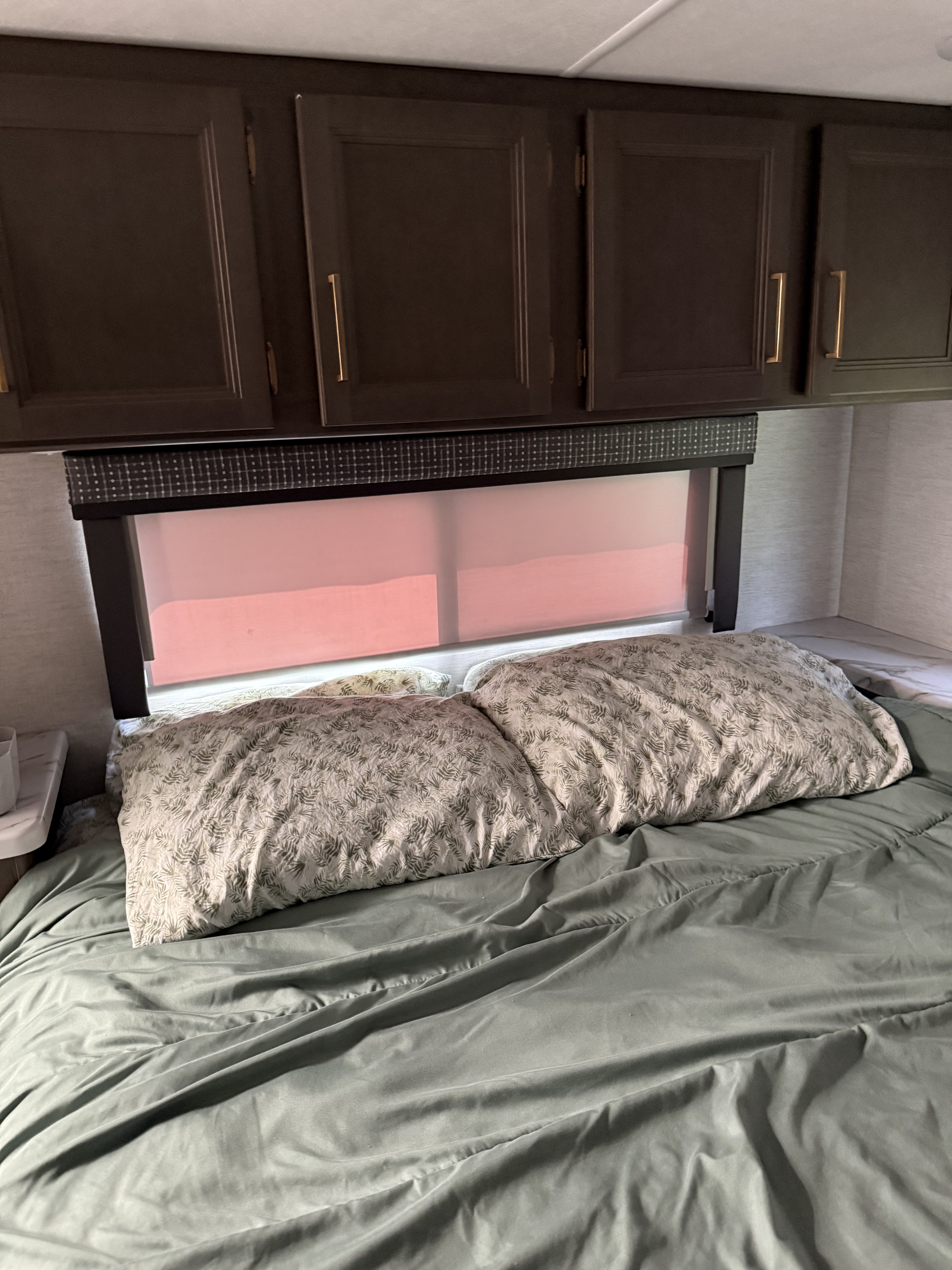 Cozy RV rental bedroom showing a bed with olive green sheets, botanical pillows, dark cabinets, and a pink window shade.. Thor Chateau 2025