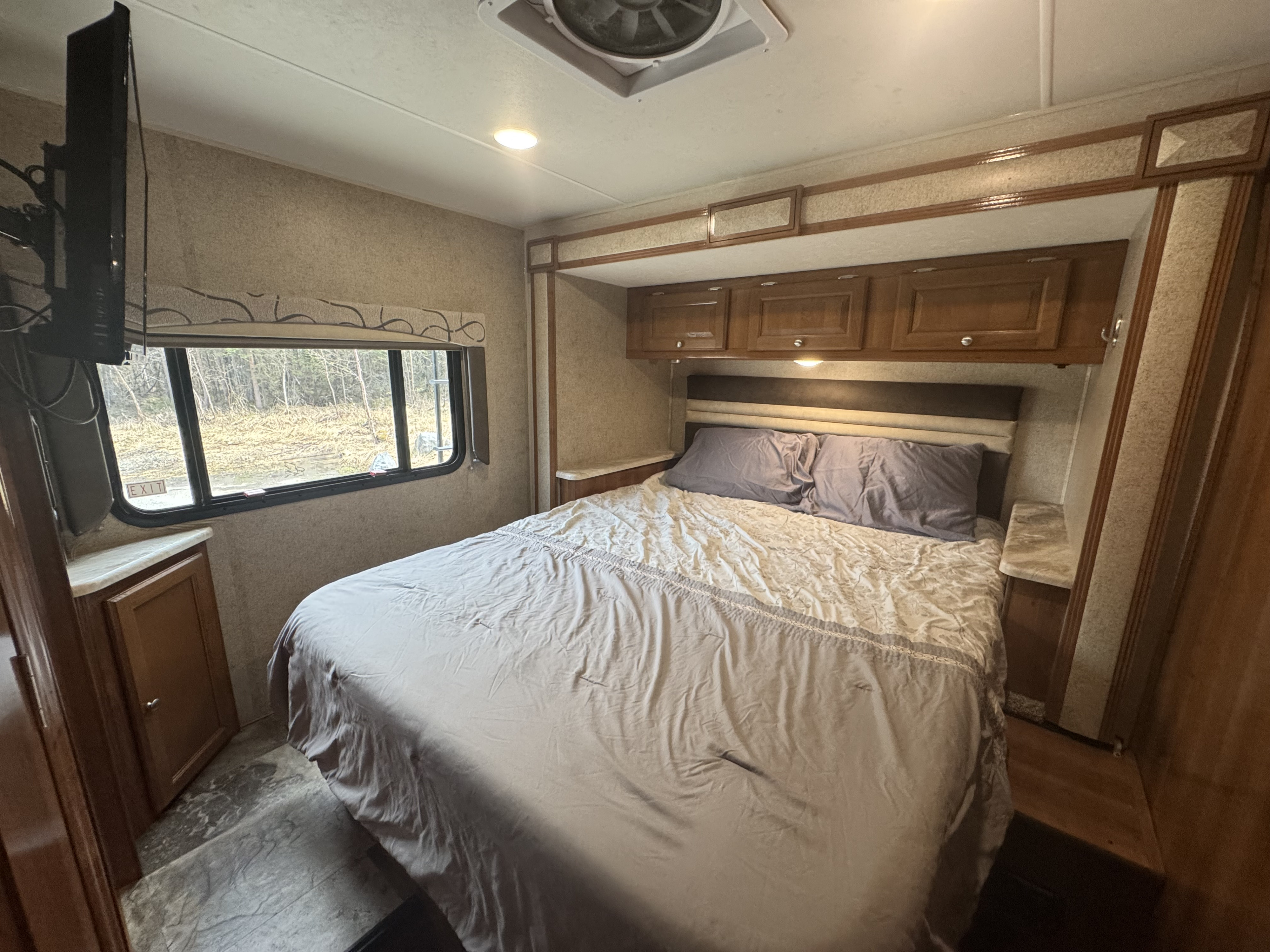 RV rental bedroom with a comfy bed, overhead cabinets, wall-mounted TV, and a window showcasing a natural outdoor view.. Coachmen Leprechaun 260DS Ford 450 2019