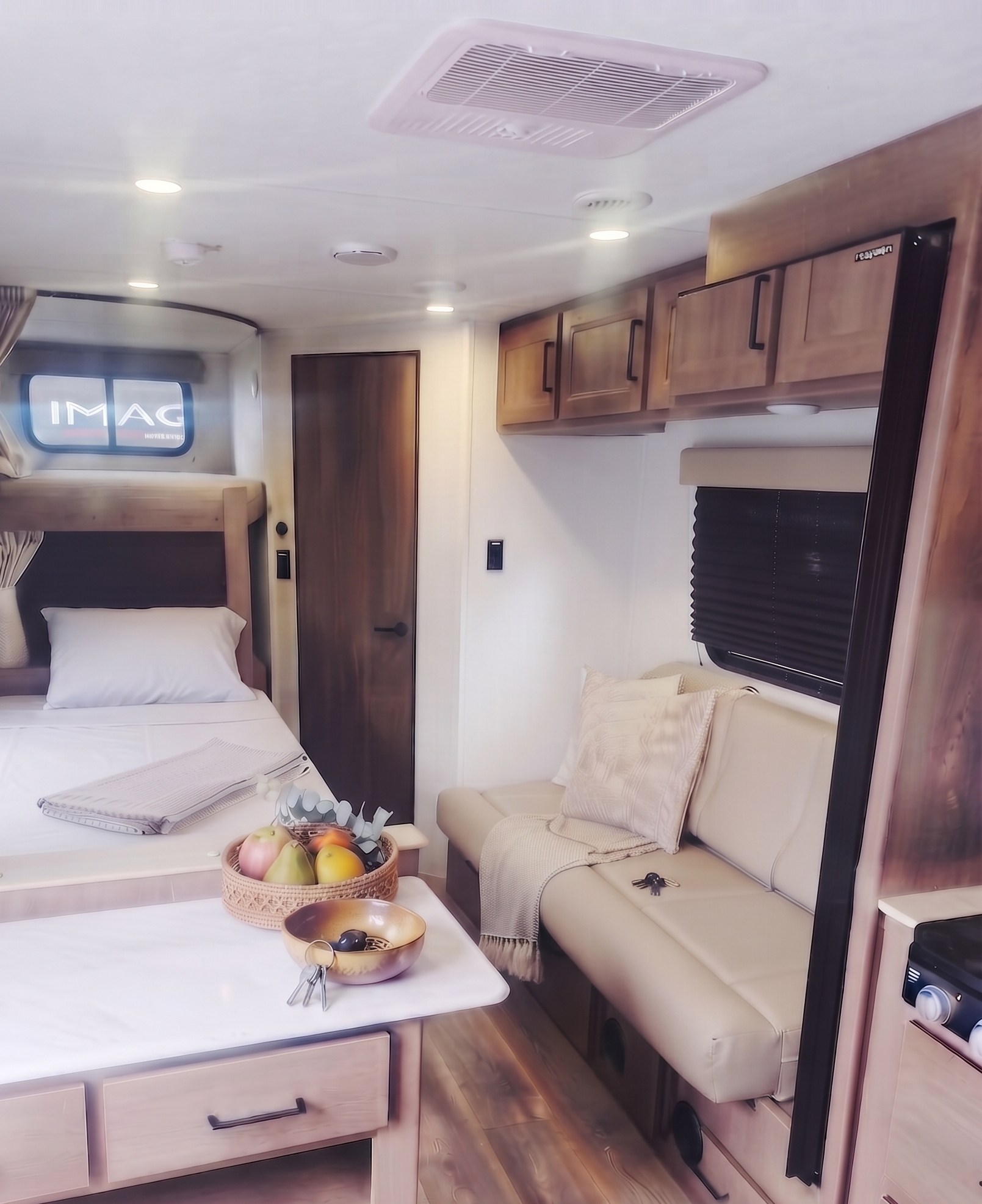 Cozy RV rental interior: a bunk bed with white linen, a light-colored sofa, and a table with fruit and keys.. Grand Design Transcend Xplor 2025