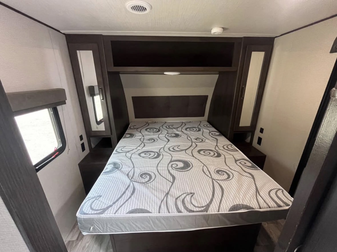 RV rental bedroom: a cozy interior with a patterned mattress, dark wood storage, and a window for natural light.. Grand Design Transcend Xplor 2022