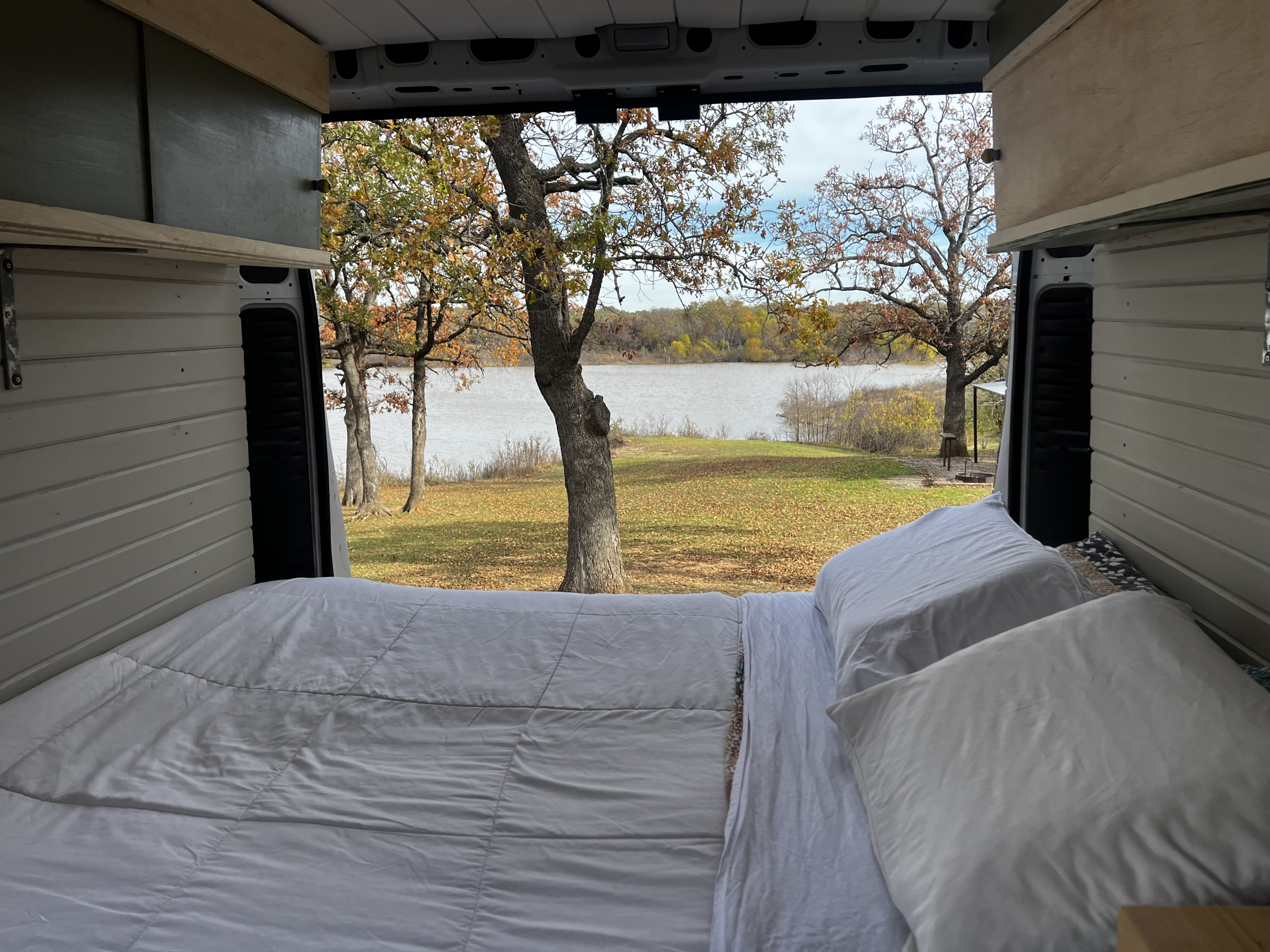 View from an RV rental bed: cozy camper van with open back doors revealing a beautiful lake and autumn trees.. Dodge Promaster 2500 high roof 2019