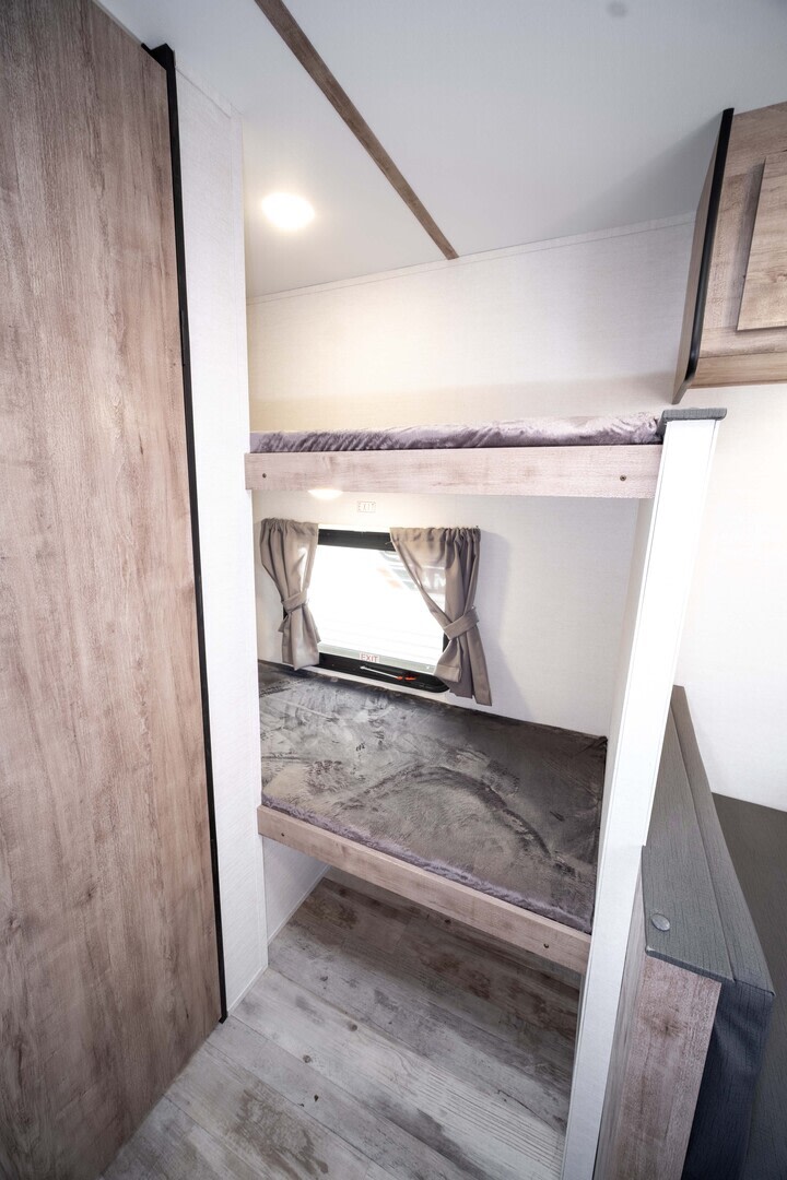 Interior of an RV rental showing modern bunk beds with gray blankets, a window, and wood-look flooring. A comfortable camper space.. Gulf Stream Enlighten Super Lite 2025