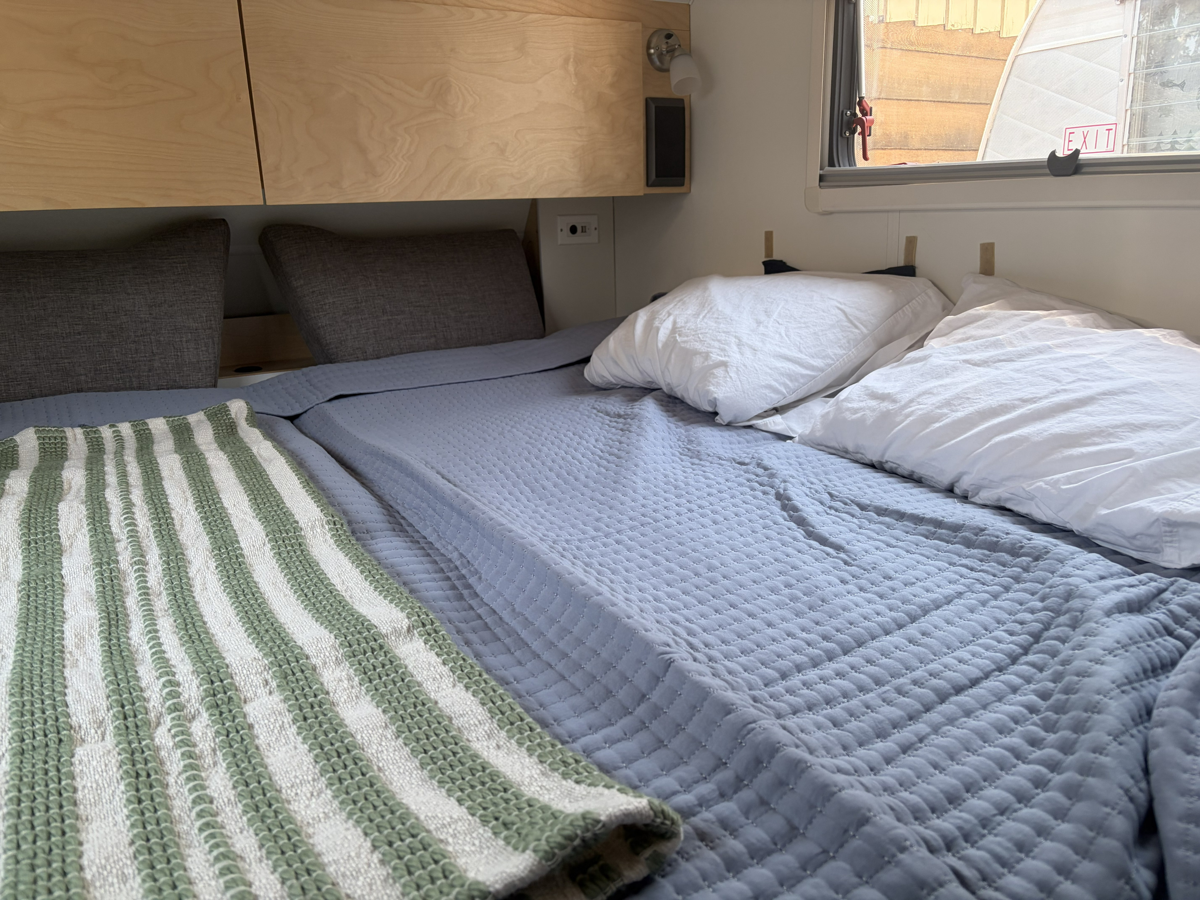 RV rental bedroom featuring a bed with quilted blue blanket, white pillows, striped throw, and wood storage.. nuCamp T@B 2021