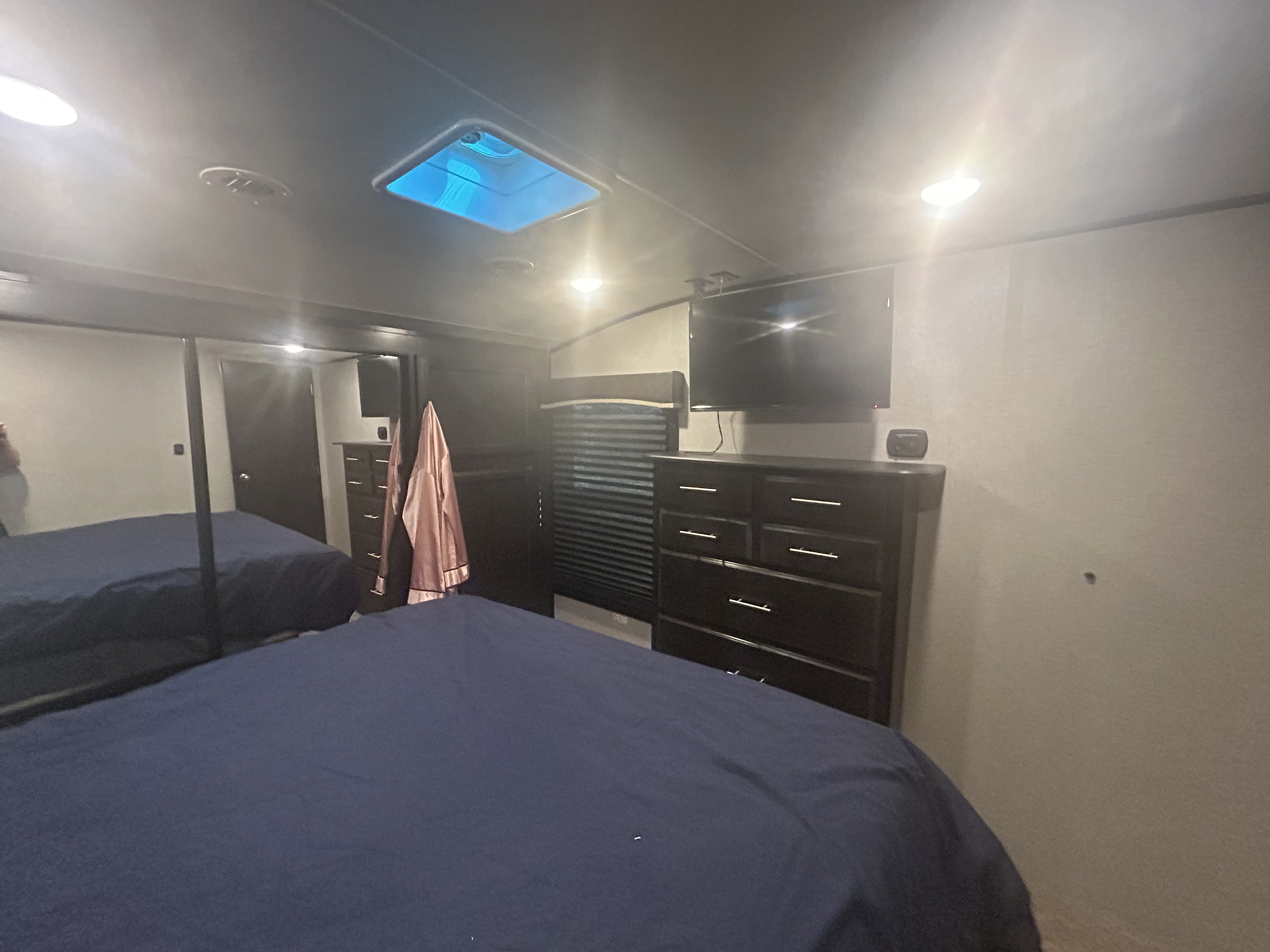 Spacious RV rental bedroom featuring a comfy navy bed, wall-mounted TV, dresser, and bright skylight. Mirror reflects more space.. Jayco Eagle 2019