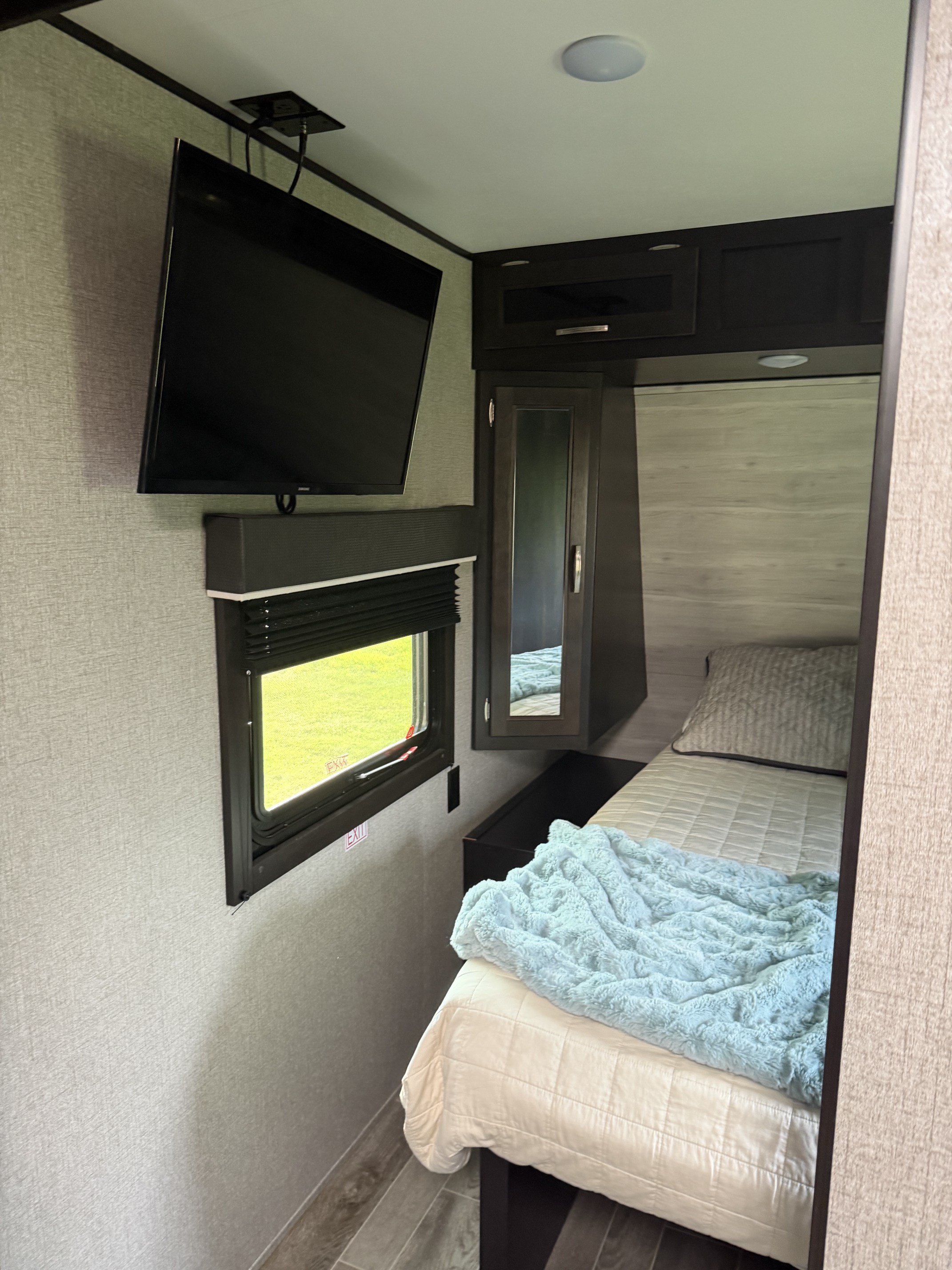 Cozy rv rental bedroom features a bed with blue blanket, wall-mounted TV, window, and dark wood storage cabinets.. Jayco Jay Flight 2021