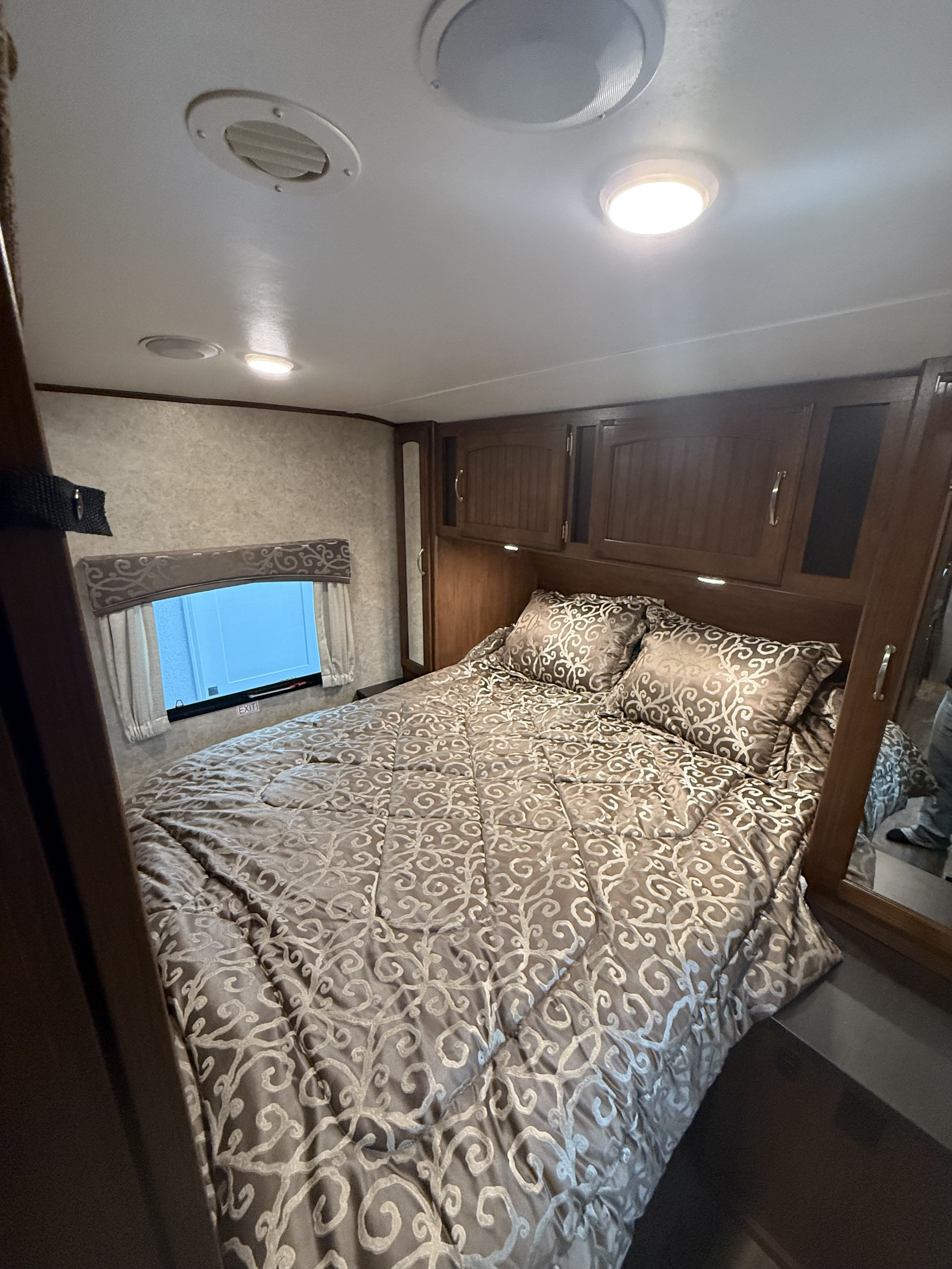 Inside a comfortable RV rental bedroom, featuring a large bed with patterned bedding, wood cabinets, and bright ceiling lights.. Jayco White Hawk 2017