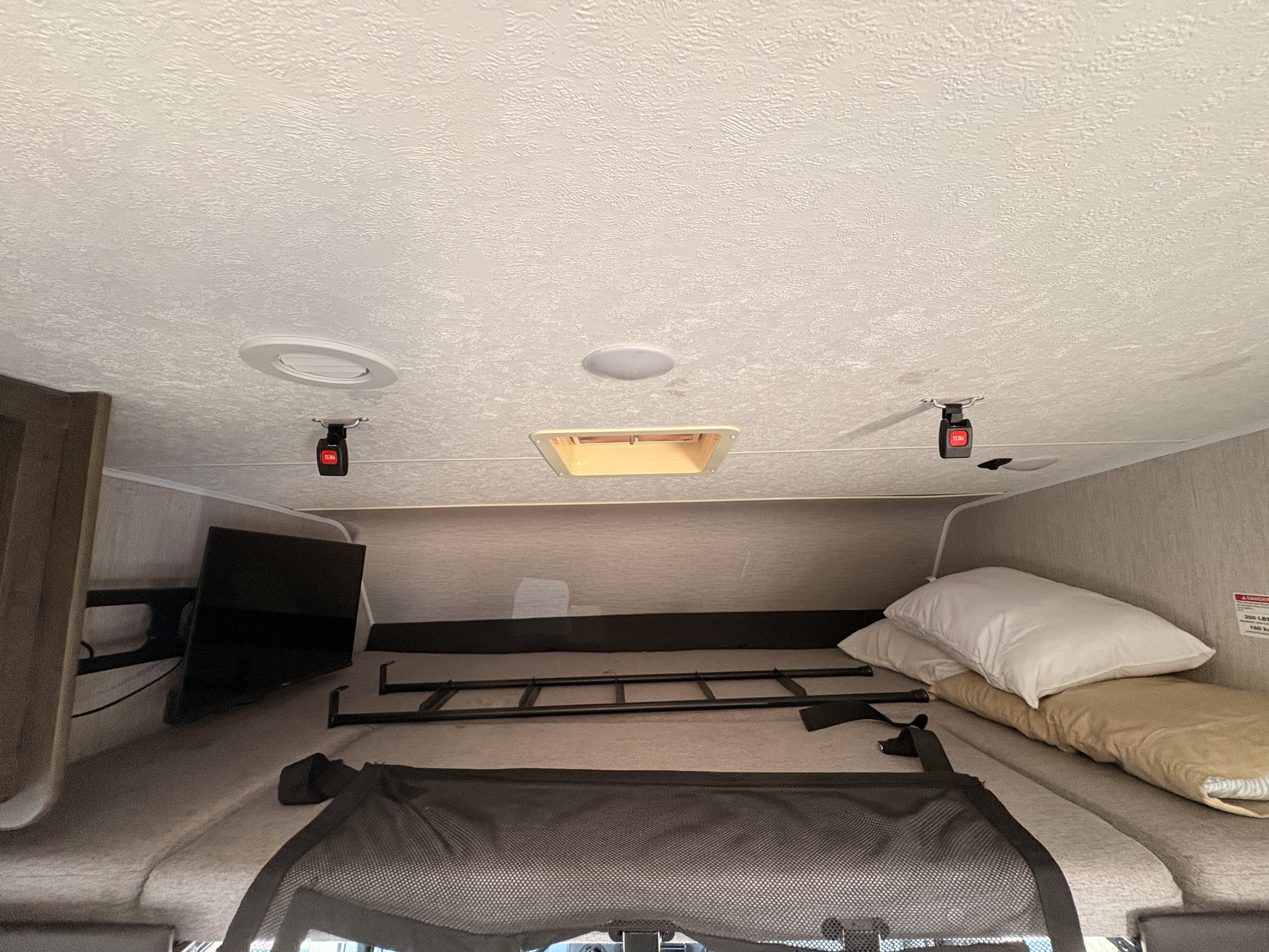 RV rental interior: cab-over bunk bed, TV, safety net, and stored ladder. Textured ceiling with lights and a vent visible.. Forest River Coachmen Leprechaun 2021