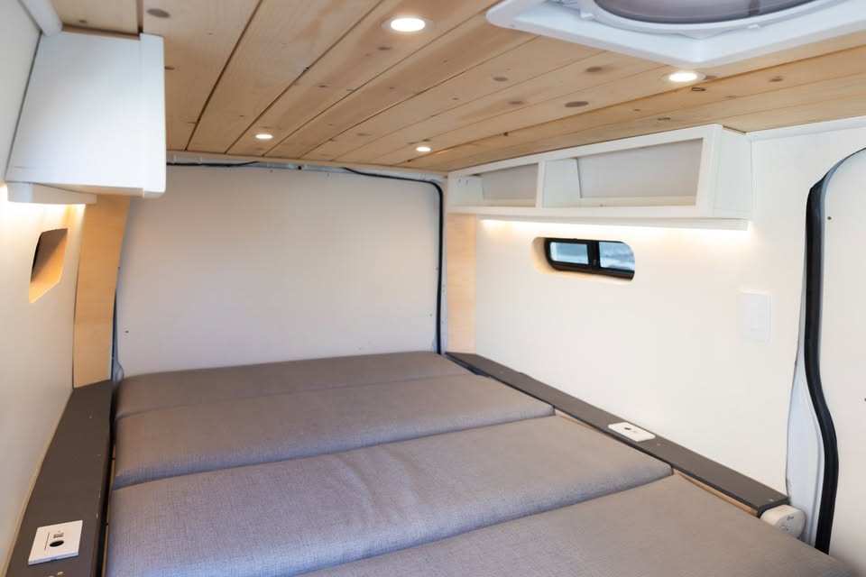 Inside an RV rental, a cozy bed with grey cushions, wooden ceiling with recessed lights, and illuminated walls, offering comfort.. Mercedes-Benz Metris Turbo 2021