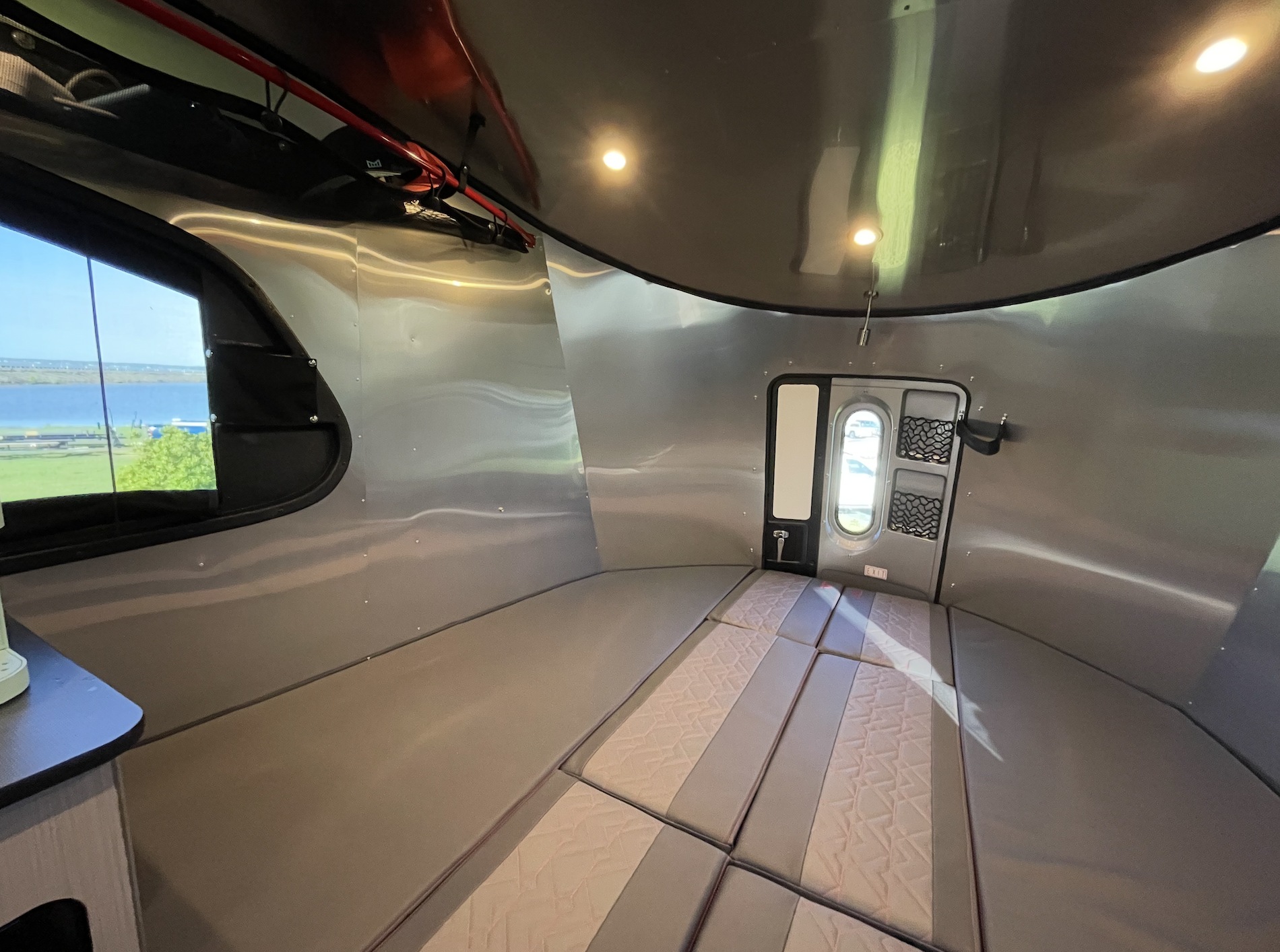 Inside a sleek rv rental: curved silver walls, a comfortable convertible bed, and a window with a water view.. Airstream Basemamp 2025