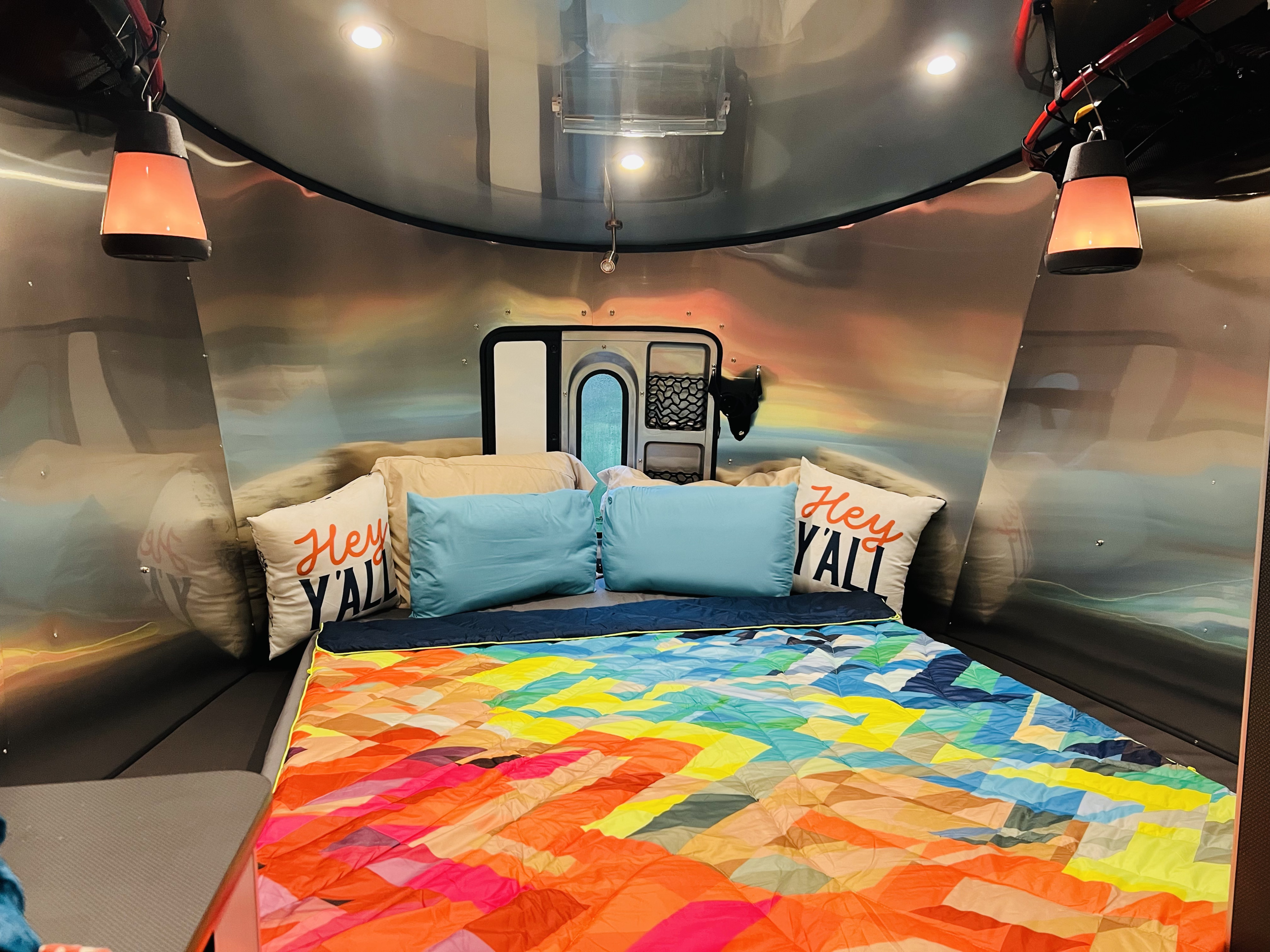 Stylish RV rental bedroom with a colorful bed, "Hey Y'ALL" pillows, hanging lanterns, and curved metallic walls.. Airstream Basemamp 2025