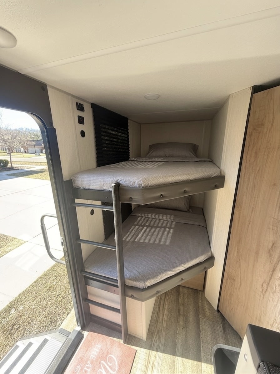 RV rental interior showcasing compact bunk beds with grey bedding and a ladder. An open door reveals the outdoors.. Forest River Campsite Reserve 2025