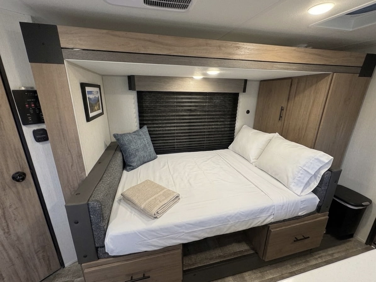 Inside a modern RV rental bedroom: comfortable bed with white linens, storage, and RV control panel for travelers.. Forest River Campsite Reserve 2025