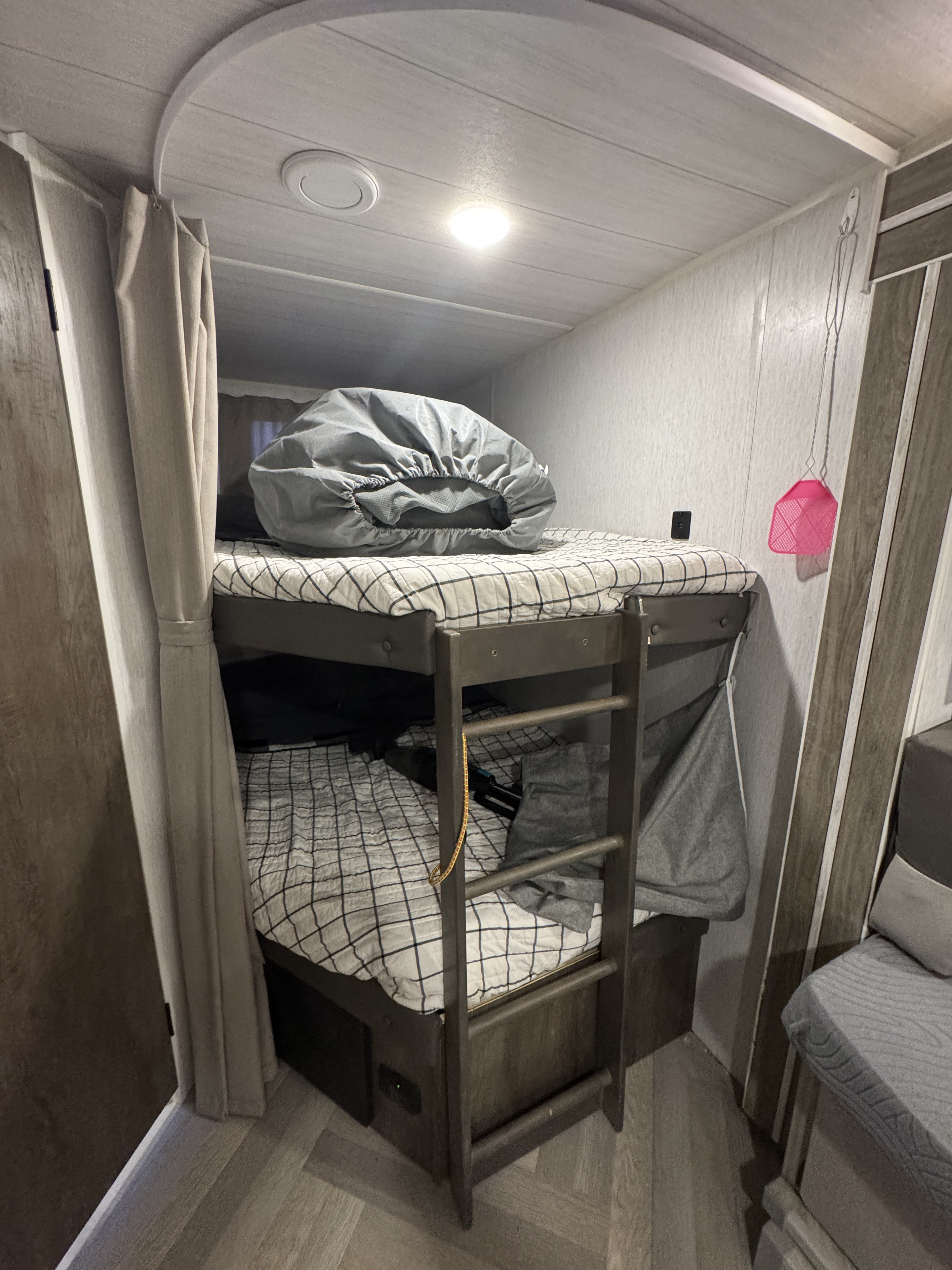 Inside an RV rental, showing a well-lit bunk bed area with a ladder, checkered mattresses, and privacy curtain.. Forest River Wildwood X-Lite 2022