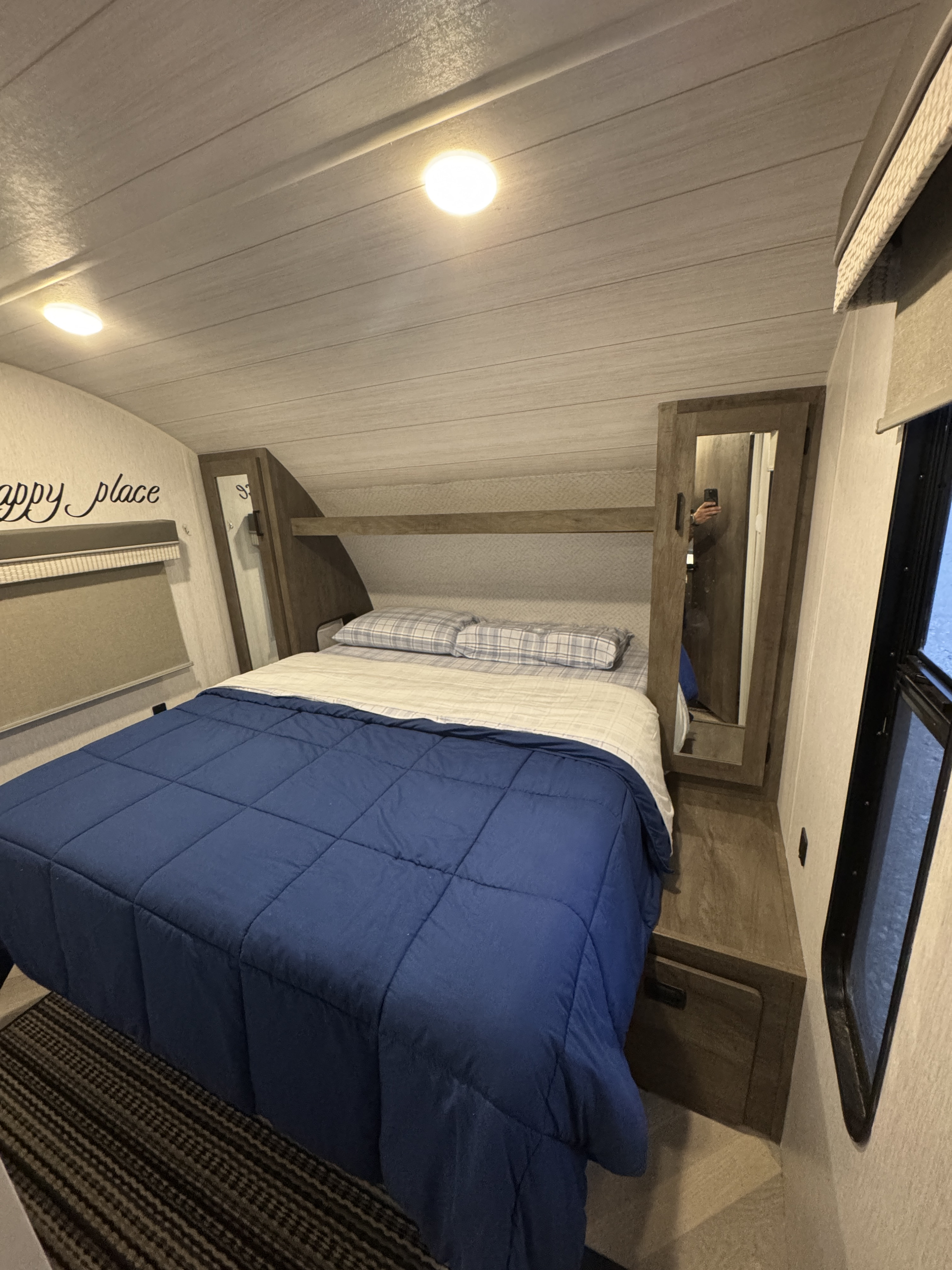 Cozy RV rental bedroom featuring a bed with blue comforter, plaid pillows, built-in storage, a mirrored cabinet, and window.. Forest River Wildwood X-Lite 2022