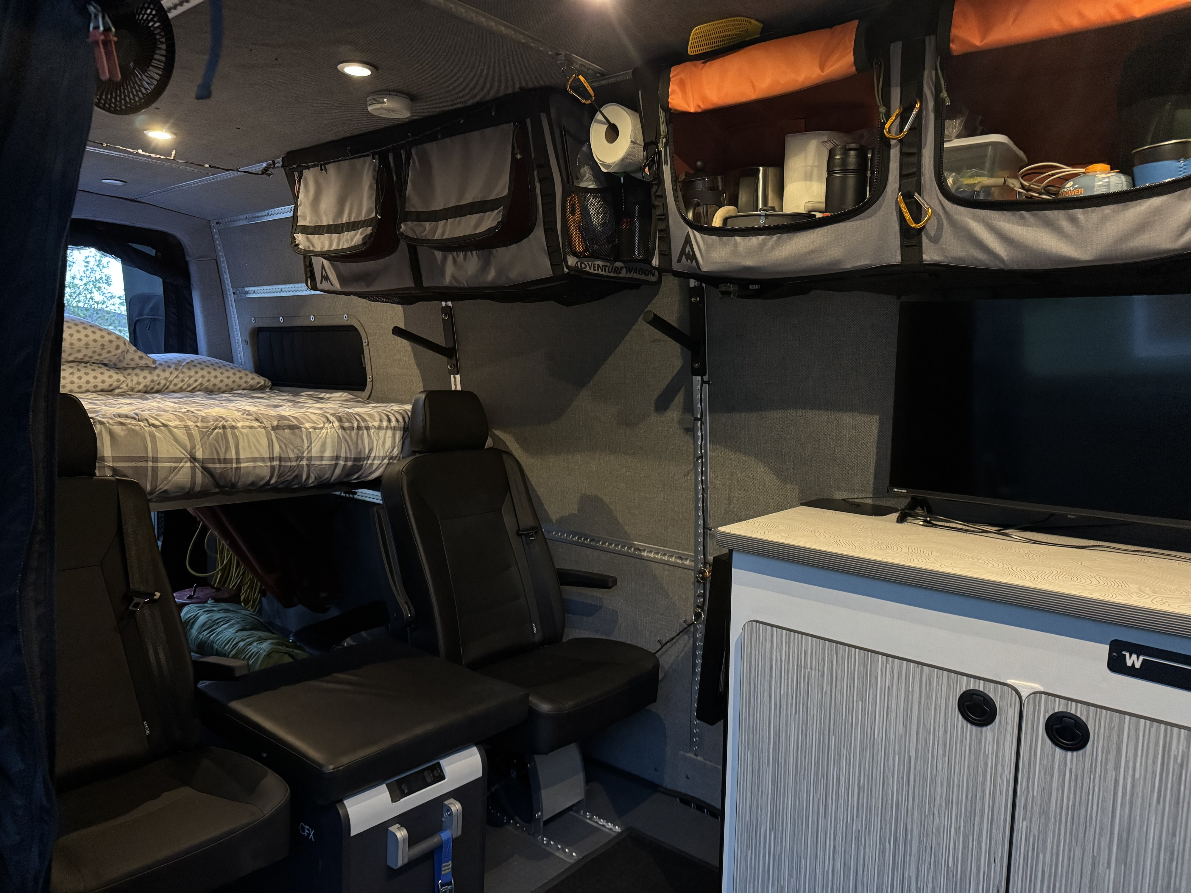 This rv rental camper van interior features a raised bed, flexible seating, overhead storage, and a counter with a TV.. Winnebago Adventure Wagon 2023