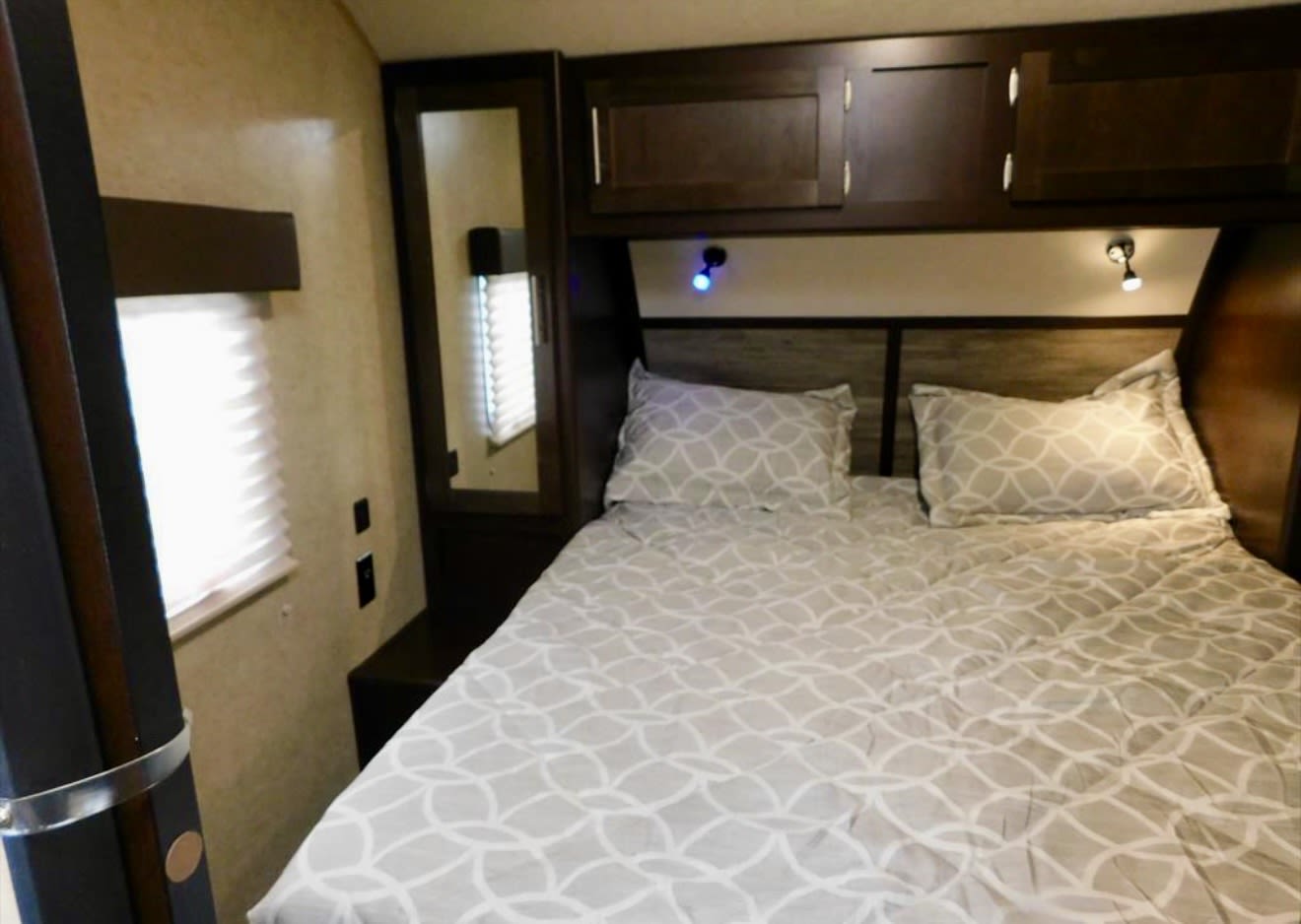 This RV rental bedroom offers a comfortable bed with patterned bedding, overhead cabinets, and a side mirror.. Forest River Cherokee 2018